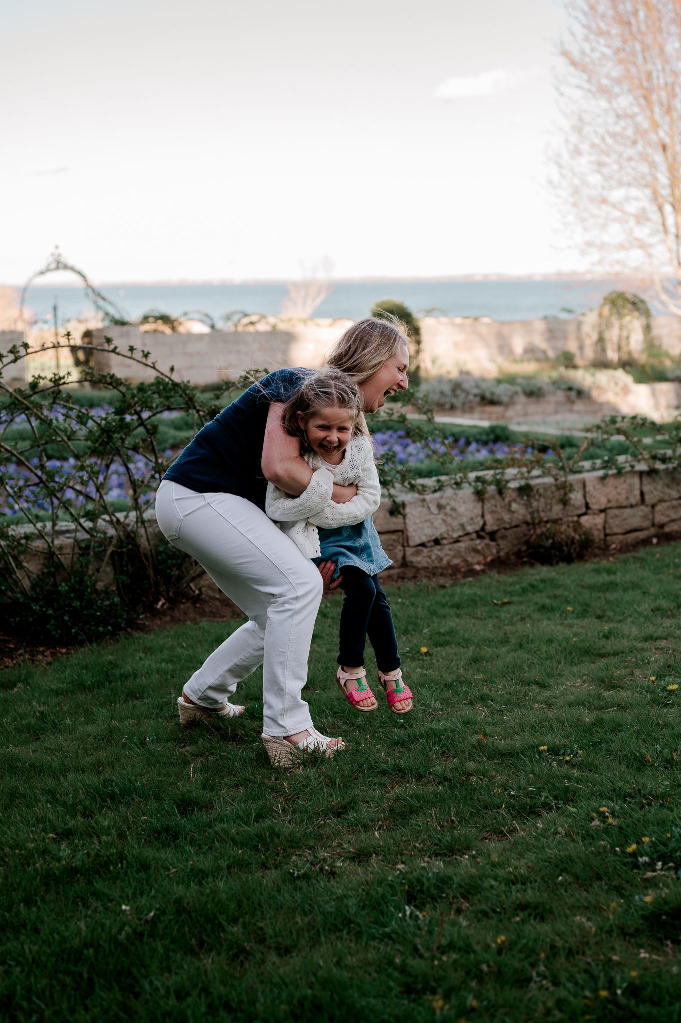 Mother joyfully lifting laughing young daughter in a garden with ocean view in background.