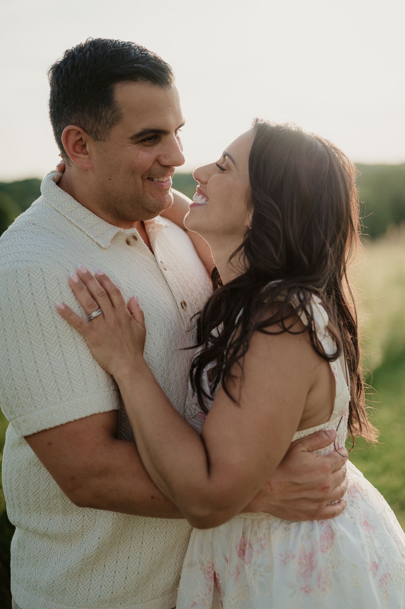 A joyful couple embraces outdoors at golden hour, the woman laughing as they hold each other close in a sunny field.