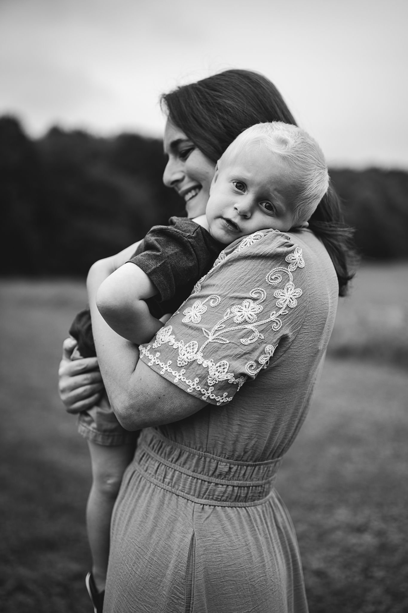 Black and white photo of a smiling mother lovingly holding her young blonde toddler outdoors in a field.