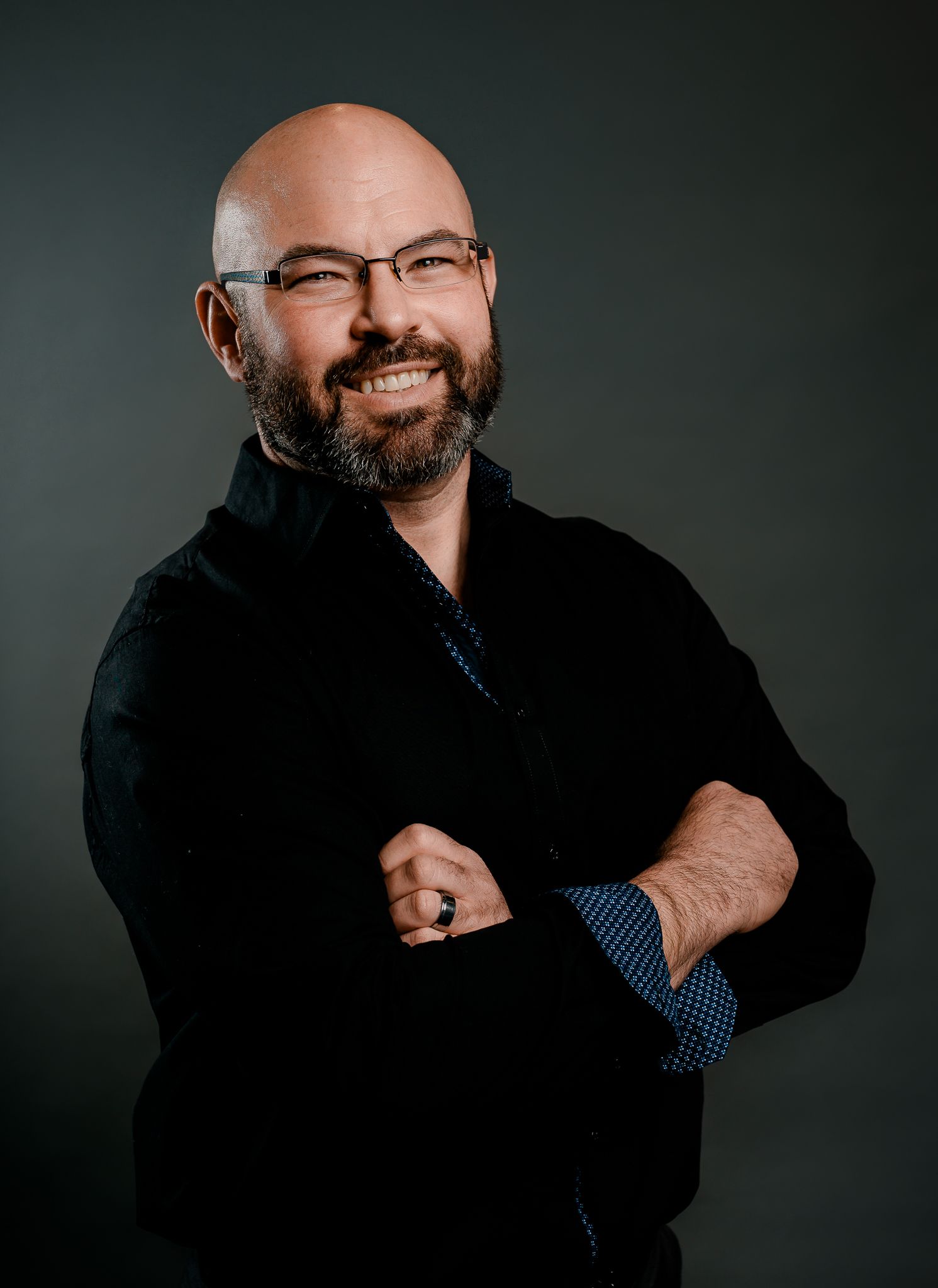 Bald bearded man in black jacket smiling with arms crossed against dark gray background.