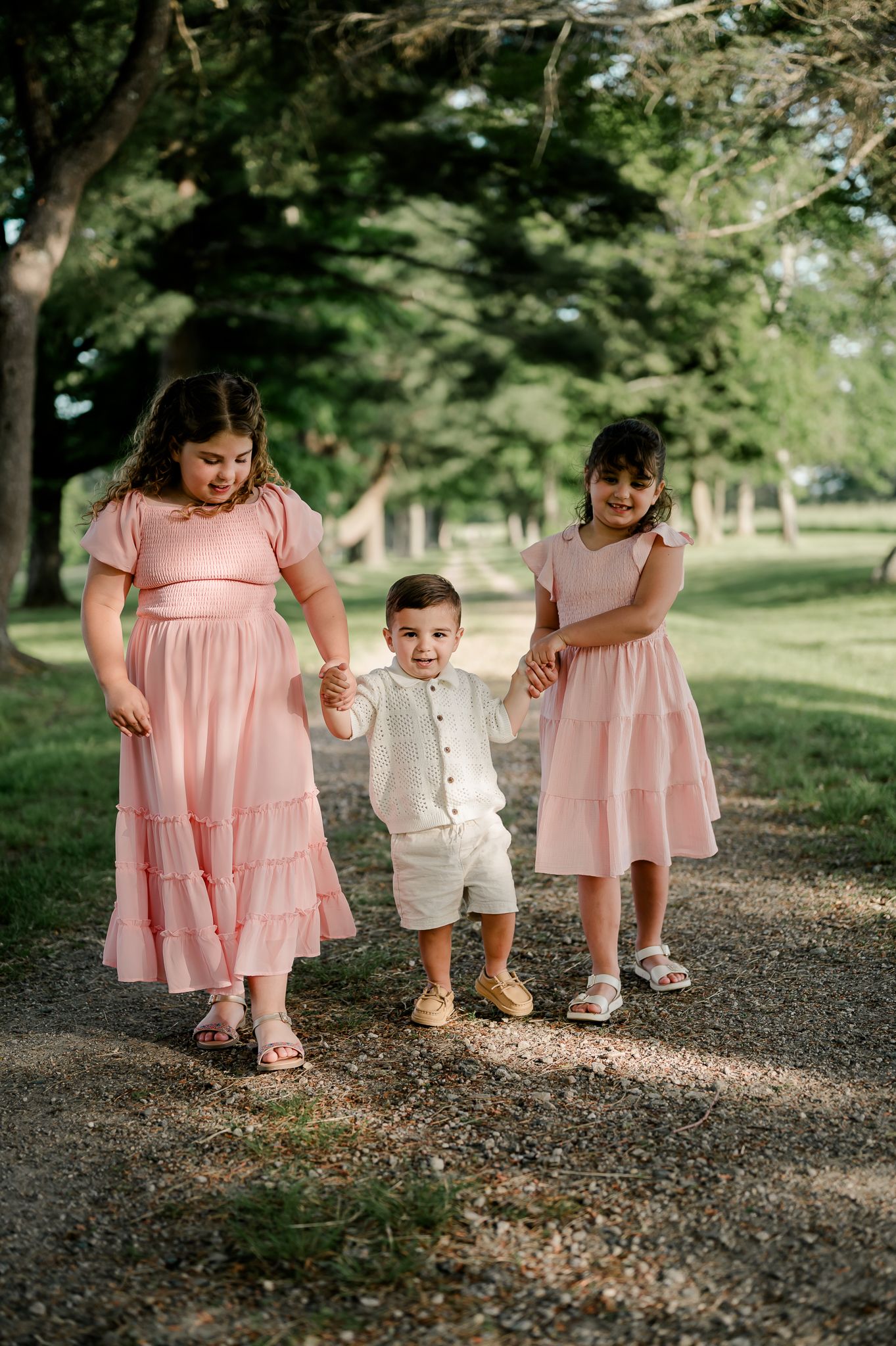 Two girls in pink dresses hold hands with a toddler boy in white on a gravel path.