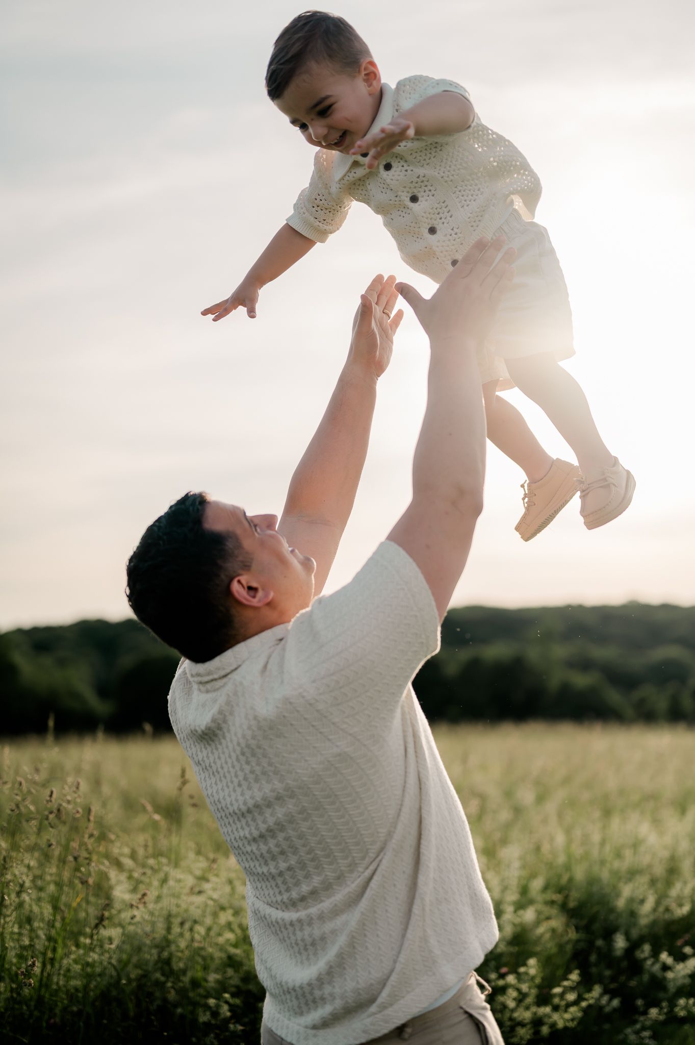 Father joyfully lifting young toddler son into the air at golden hour in a sunny meadow.