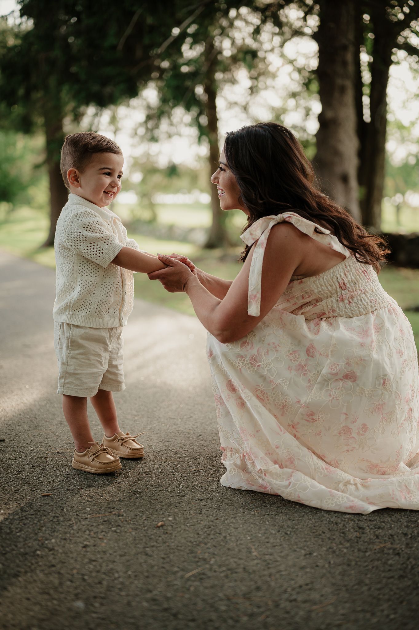 A woman in a floral dress crouches to hold hands with a smiling toddler boy on a tree-lined path.