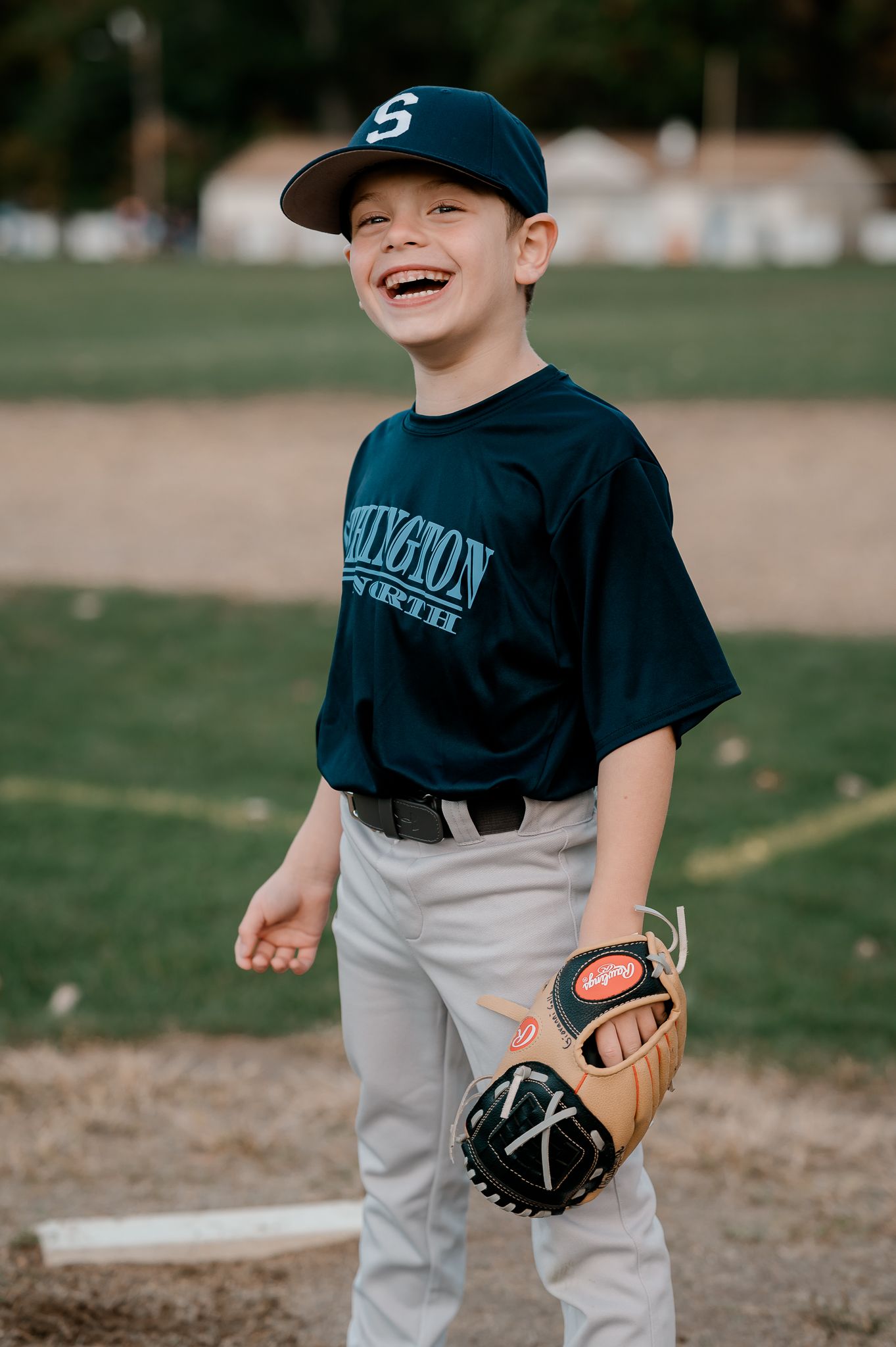 Young boy in baseball uniform laughing on field, wearing navy cap and glove.