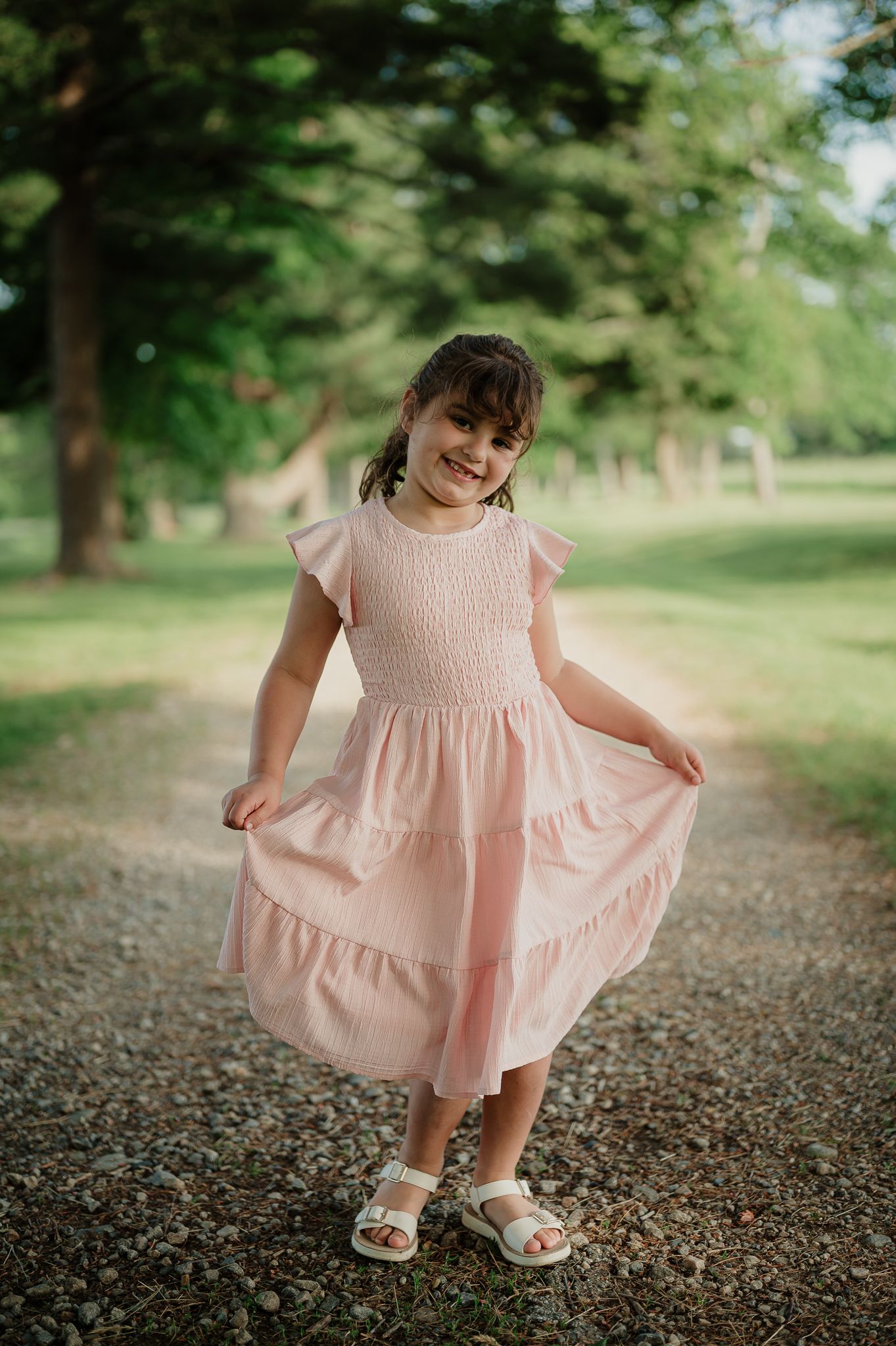 Young girl in pink smocked dress twirling on a gravel path surrounded by green trees.
