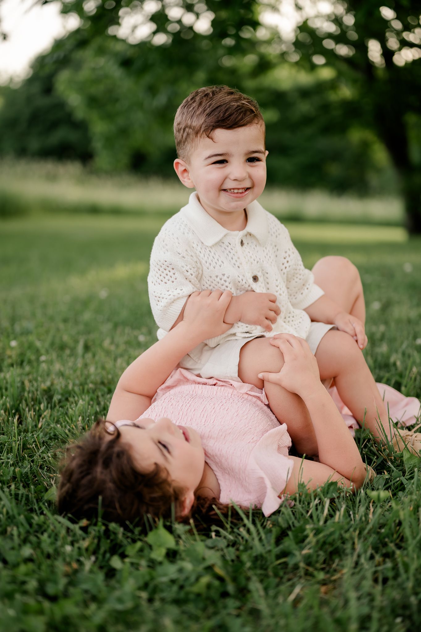 Two young siblings playing on green grass, smiling boy sitting while girl lies across his lap.