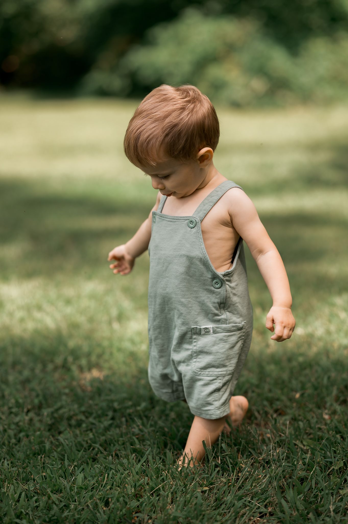 Adorable toddler in sage green overalls walking barefoot on lush green grass outdoors.