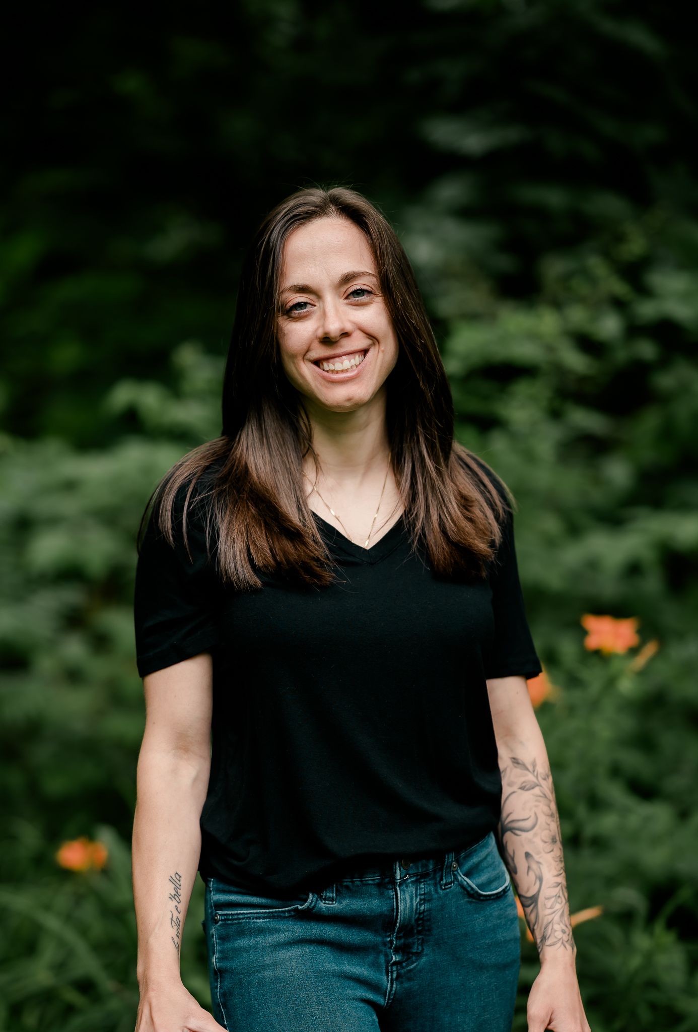 Smiling woman with tattoos wearing black t-shirt and jeans standing outdoors among green foliage.