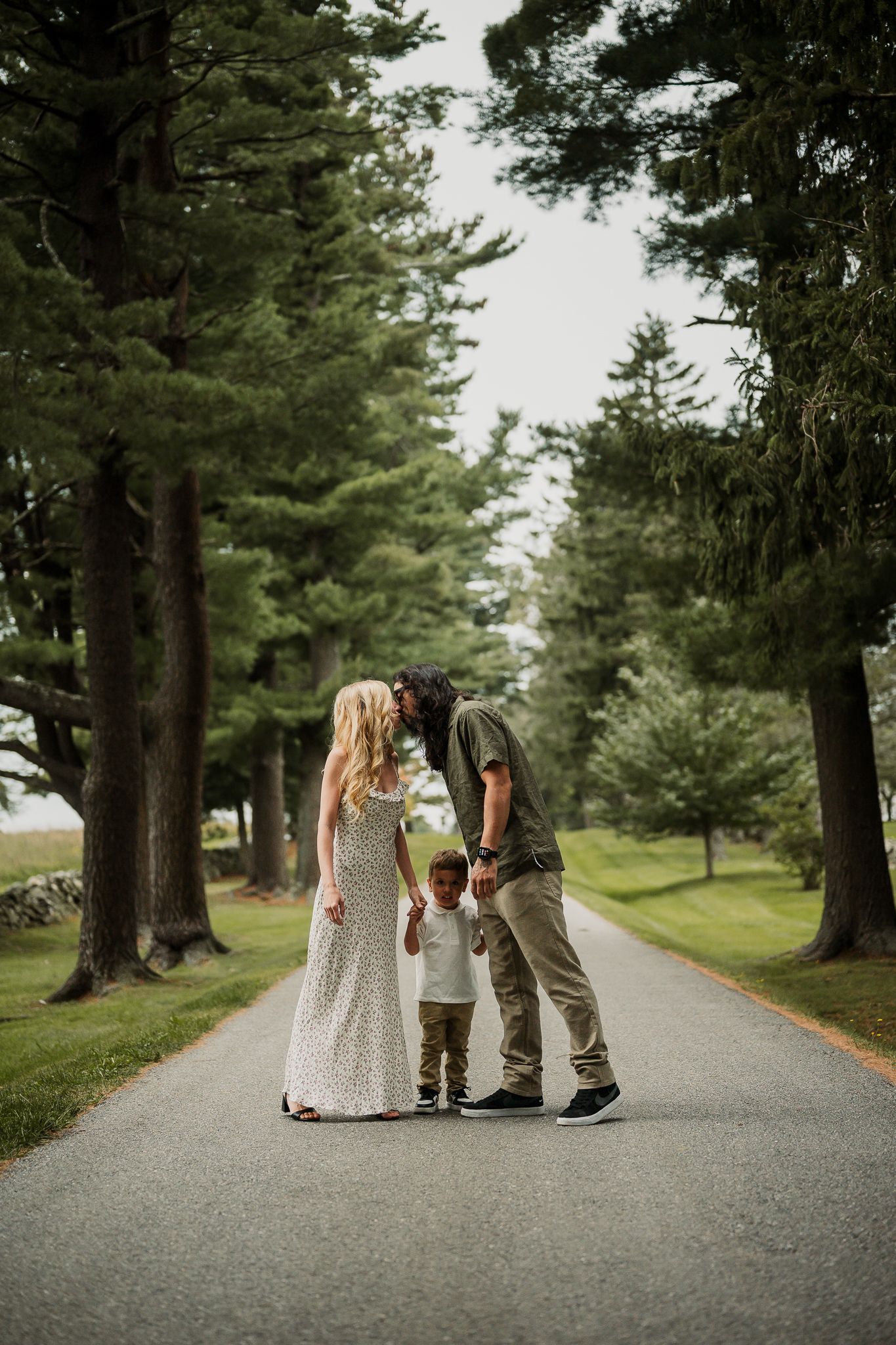 Family of three sharing a tender moment on a tree-lined path, parents leaning in to kiss above their toddler.