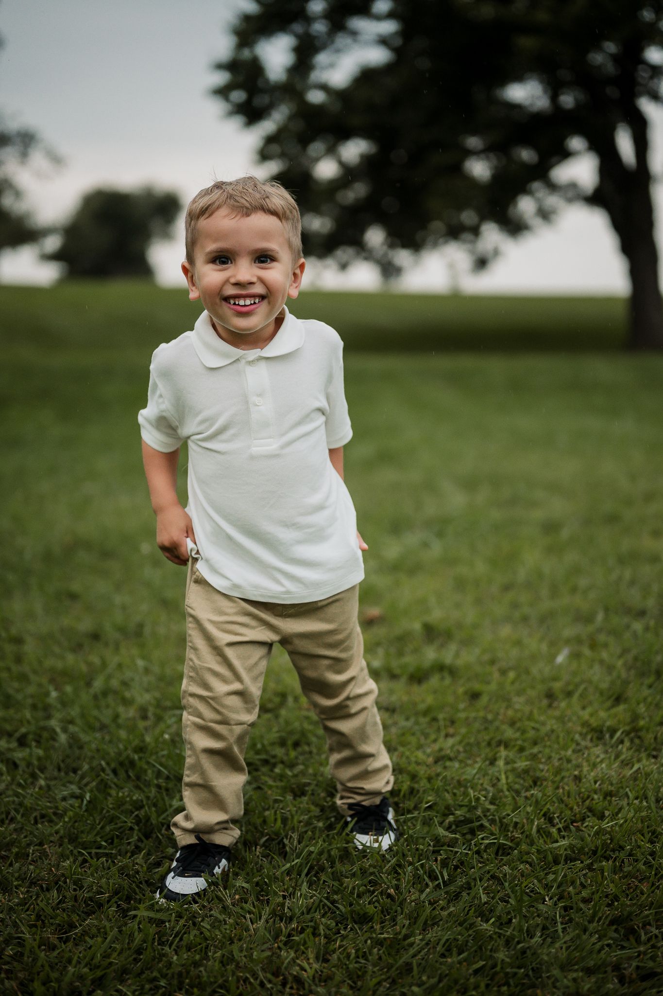 A smiling young boy in a white polo shirt and khaki pants stands on green grass outdoors.