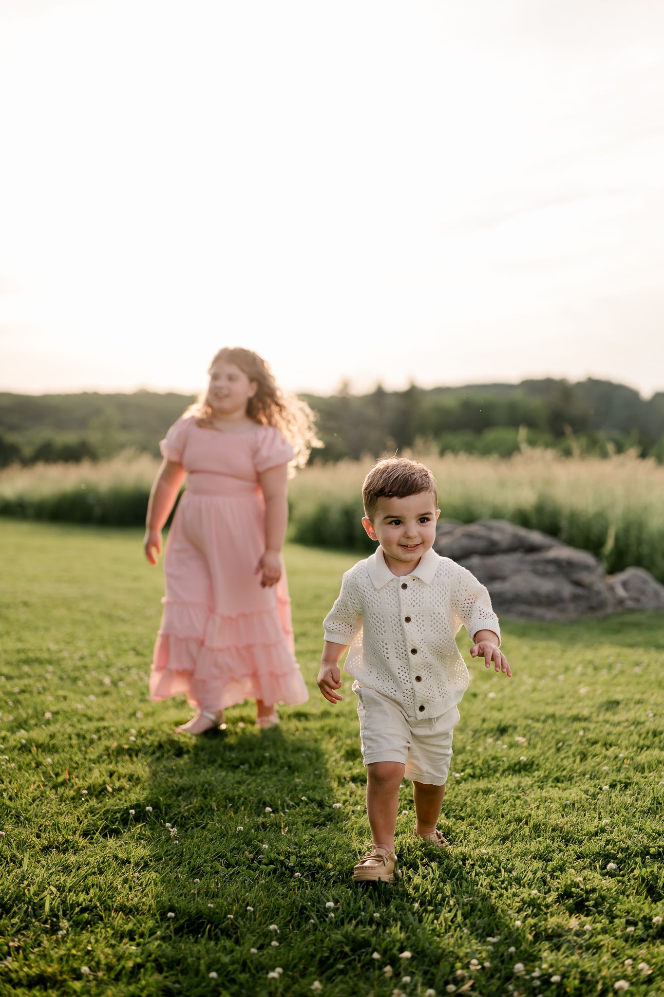 A young boy in white runs on green grass while a girl in a pink dress walks behind.