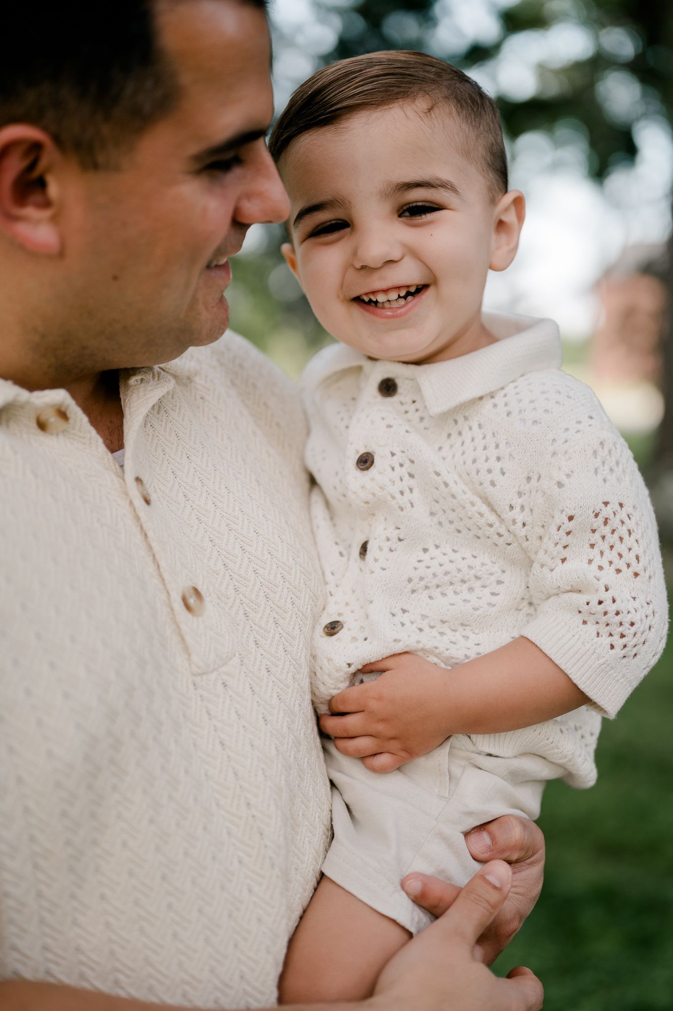 A smiling baby boy in a cream knit outfit is held by a man in a matching sweater.
