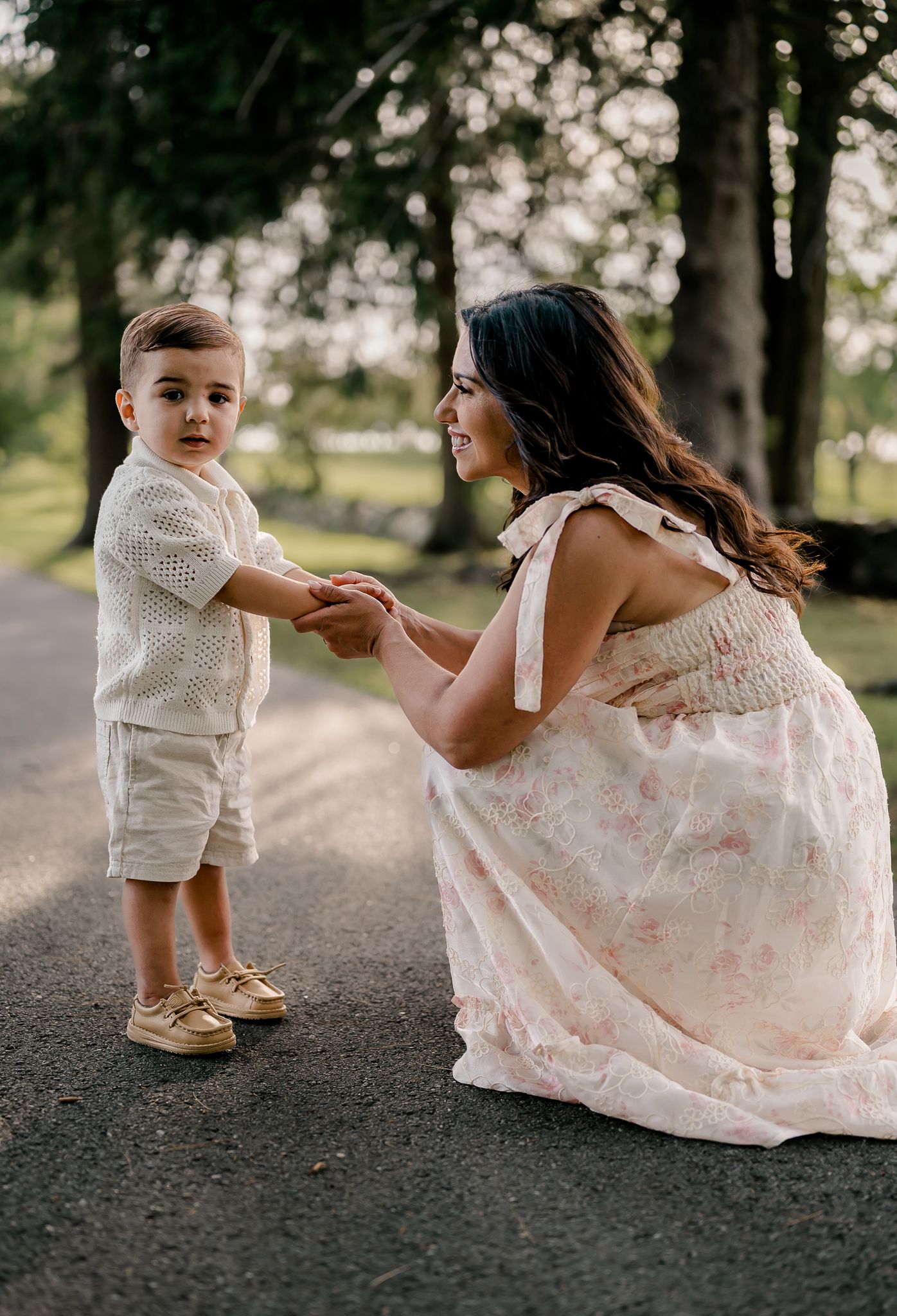 A mother in a pink floral dress kneels to hold hands with her toddler son outdoors.