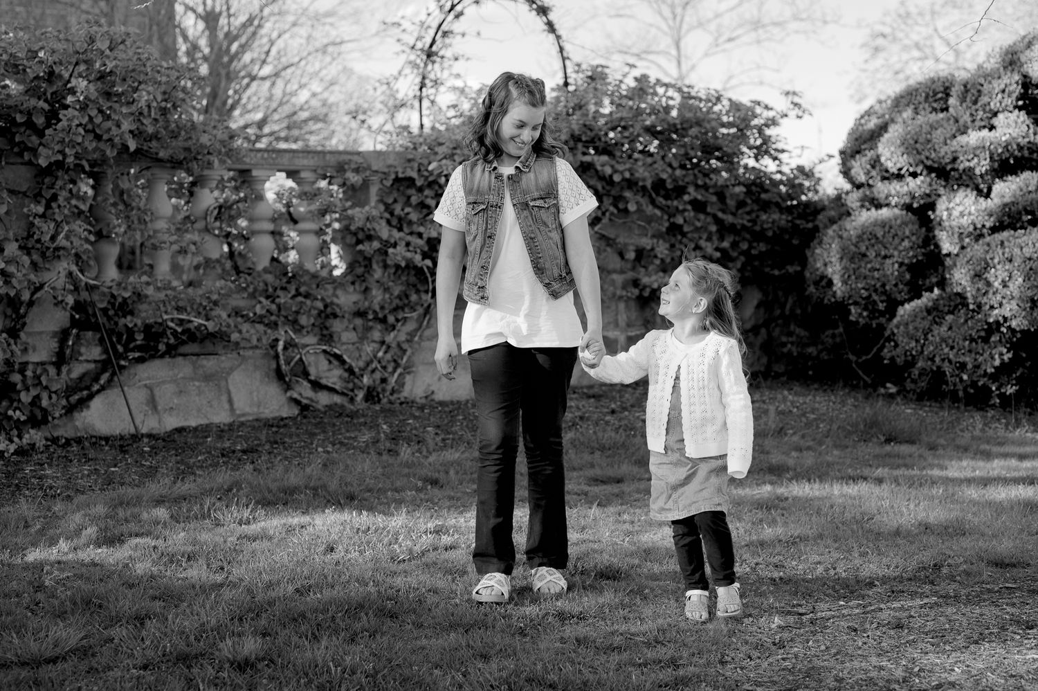 Two girls holding hands in a garden, older sister looking down at younger sibling in black and white photo.
