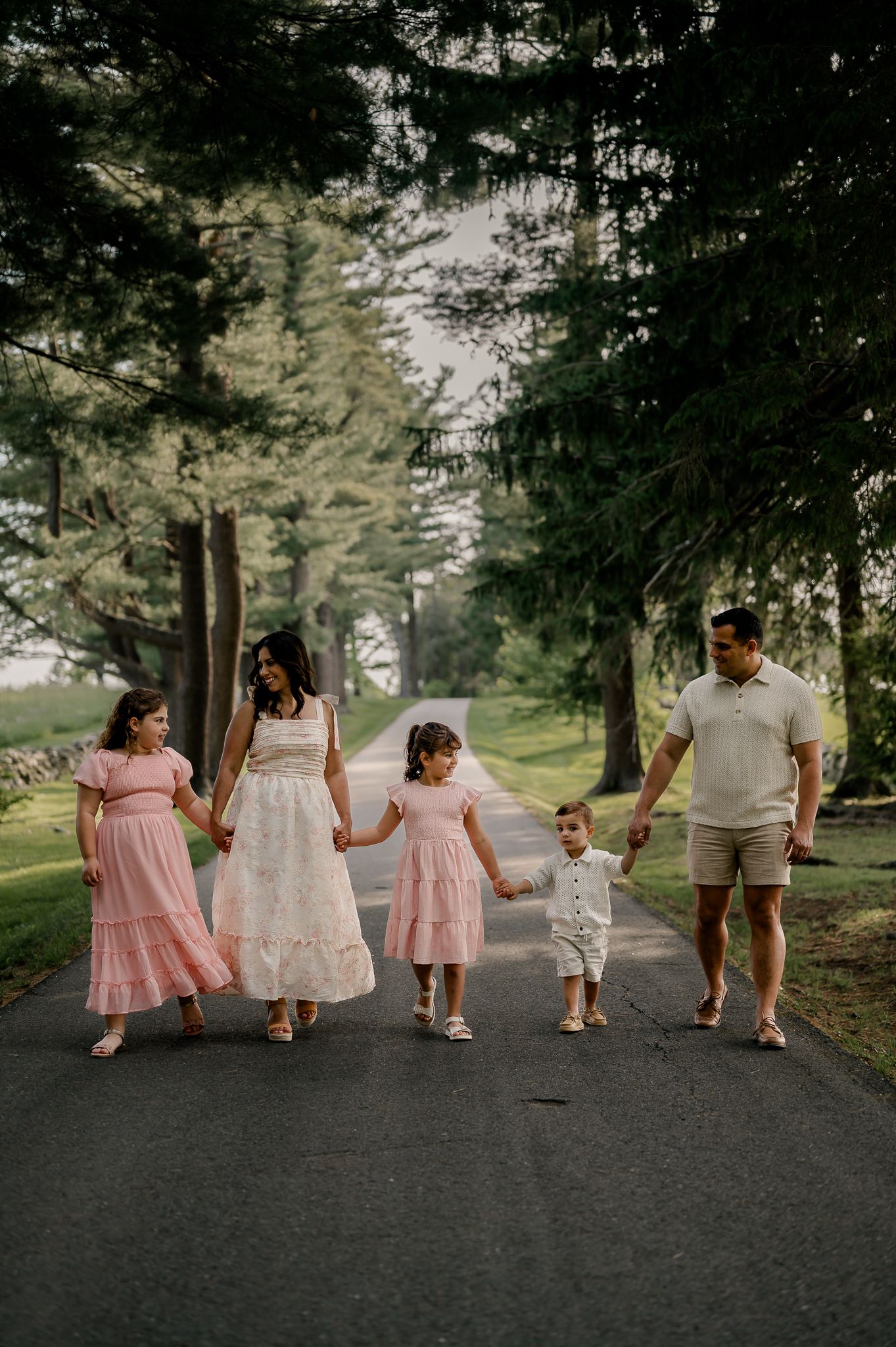 Family of five walking hand-in-hand down a tree-lined path, wearing coordinated pink and cream outfits.