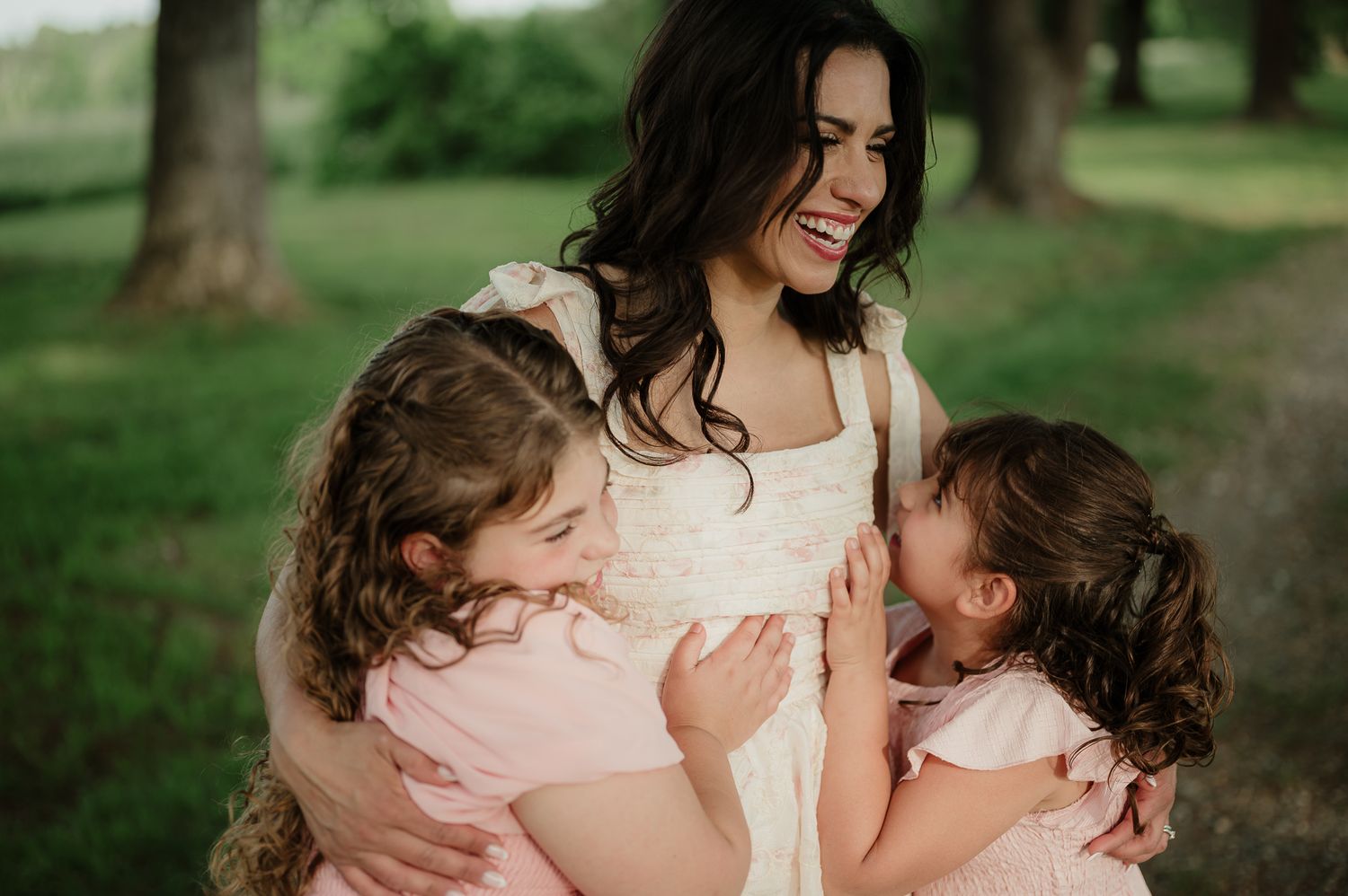 A bride in a white dress laughs joyfully as two young girls in pink dresses hug her outdoors.