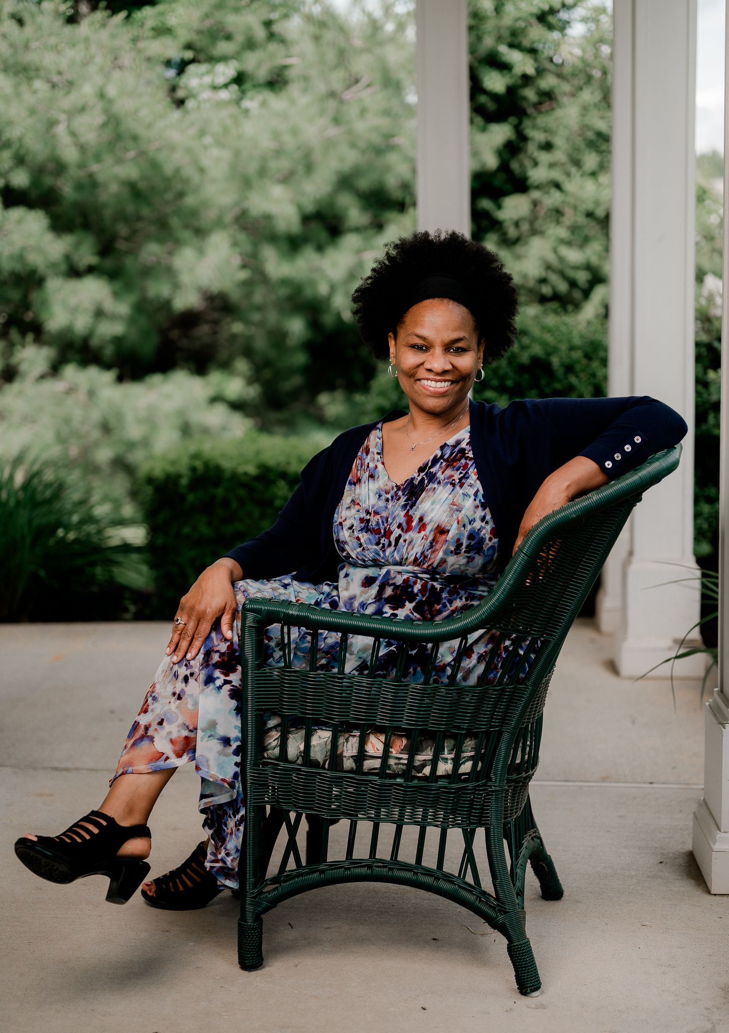 A smiling woman in a floral jumpsuit sits relaxed in a green wicker chair on a porch.