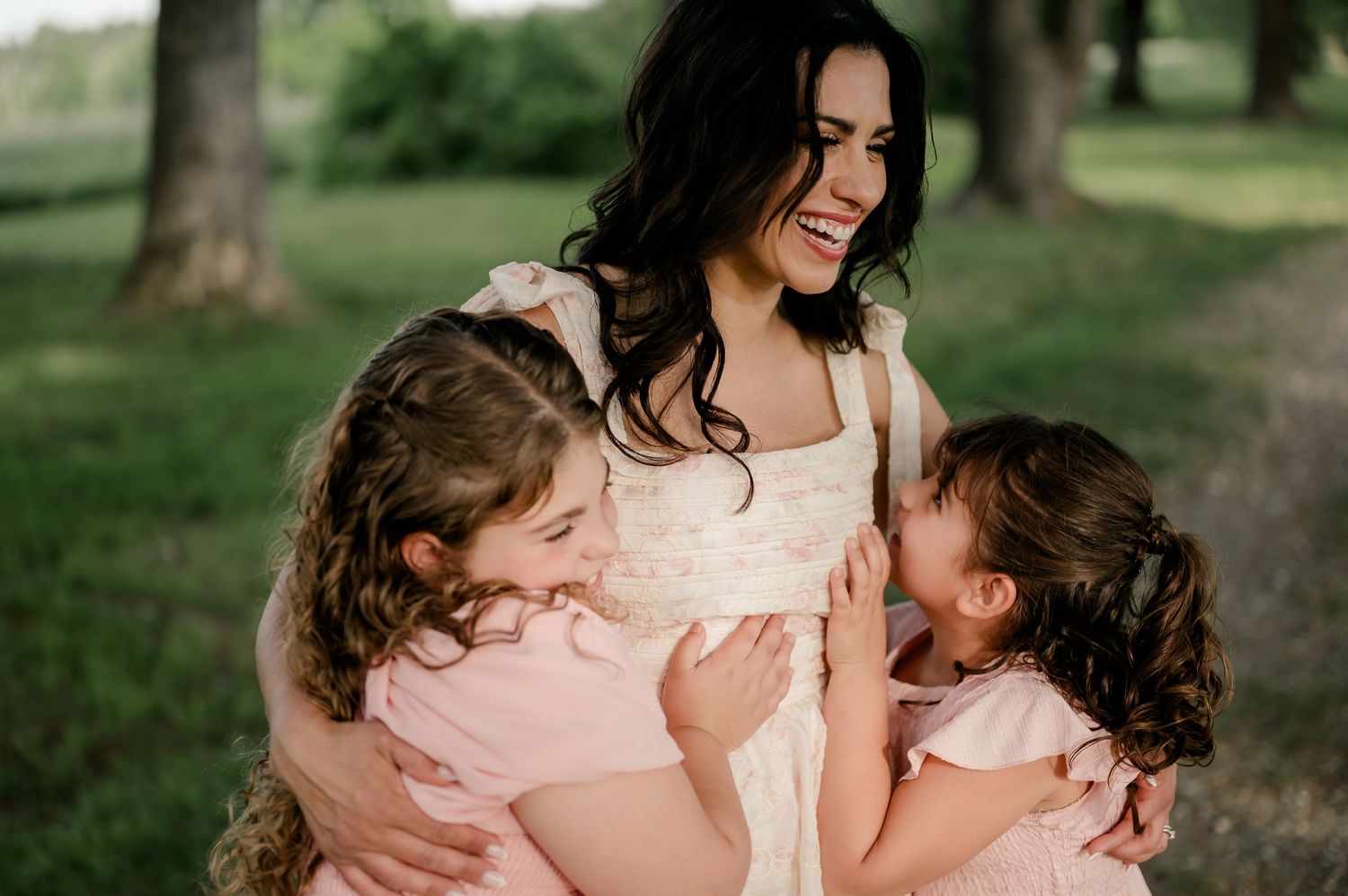Smiling dark-haired mother hugging two young daughters in matching pink dresses outdoors.