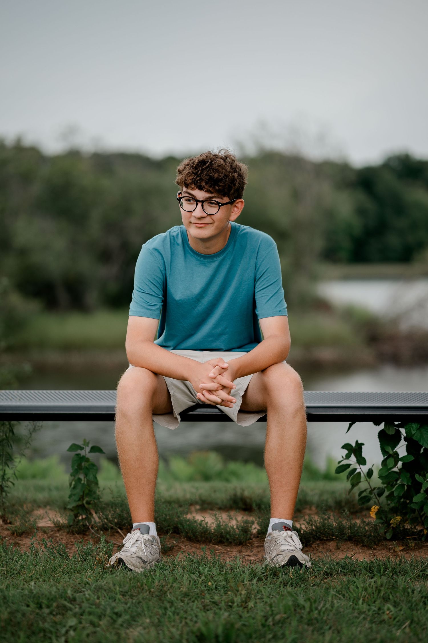 Teen boy with glasses in blue shirt sitting on bench outdoors near greenery and water.