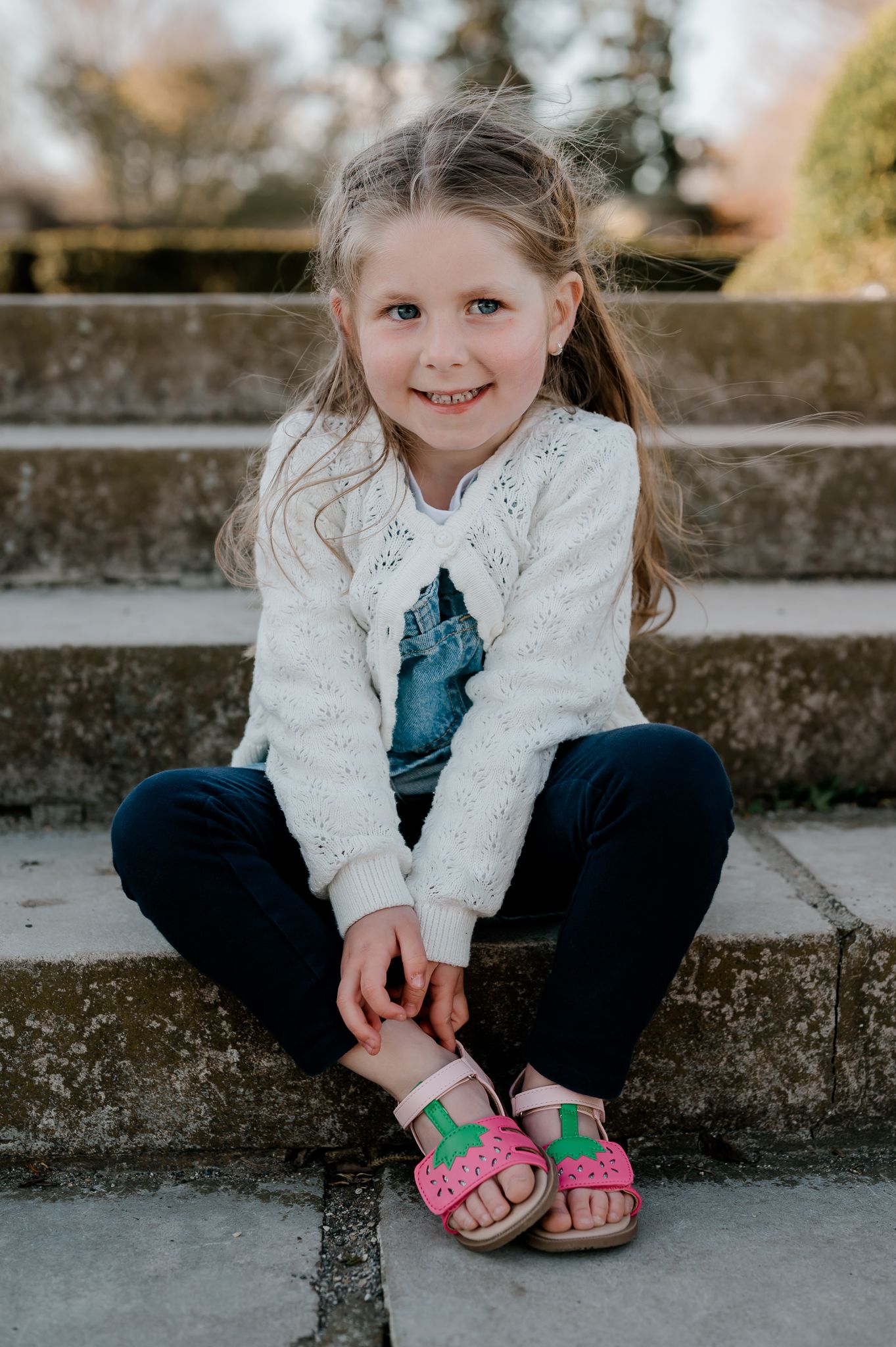 Happy young girl in white lace cardigan and pink sandals sitting on stone steps outside.