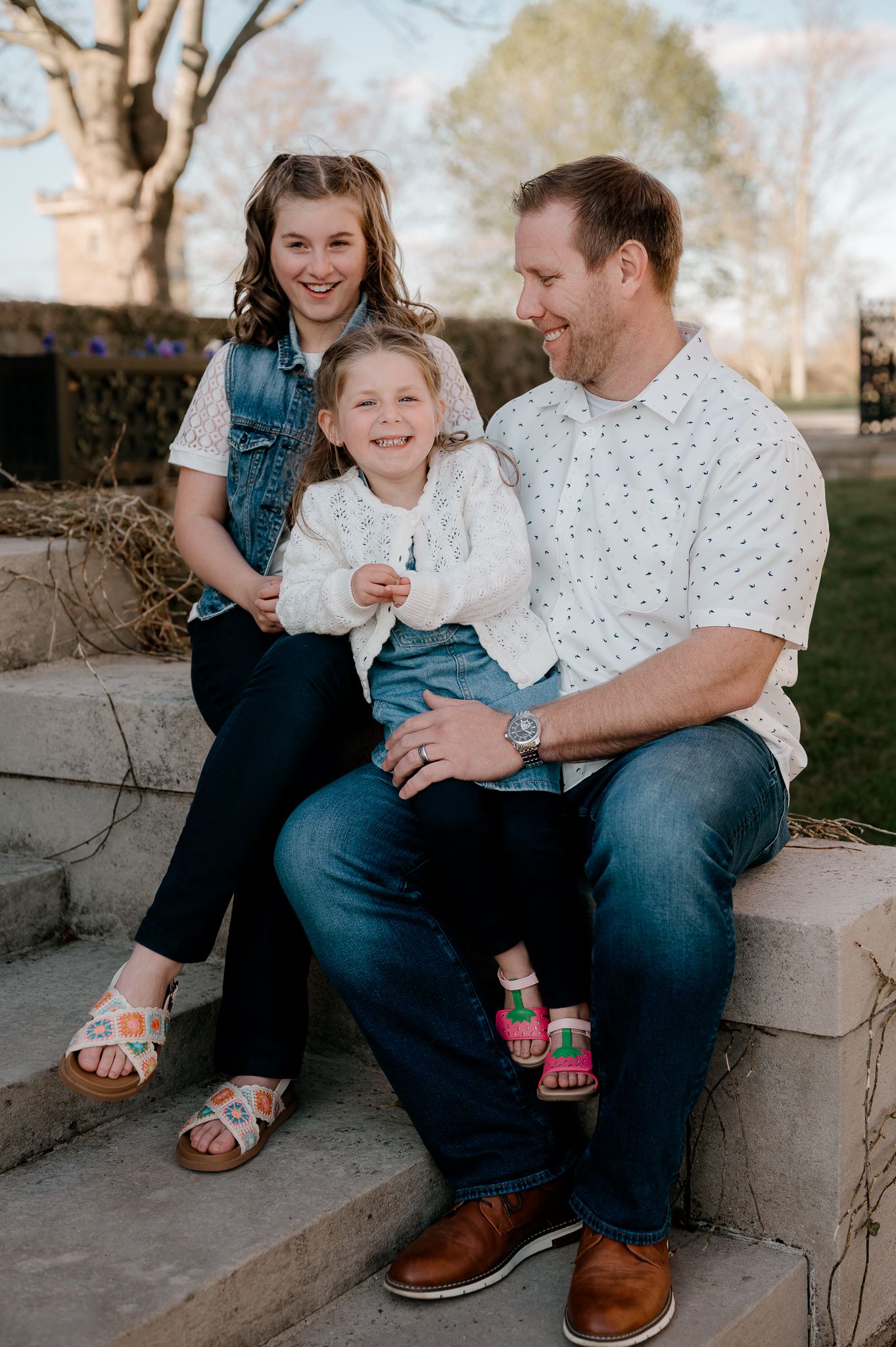 Father sitting on stone steps smiling with two daughters in outdoor spring setting.