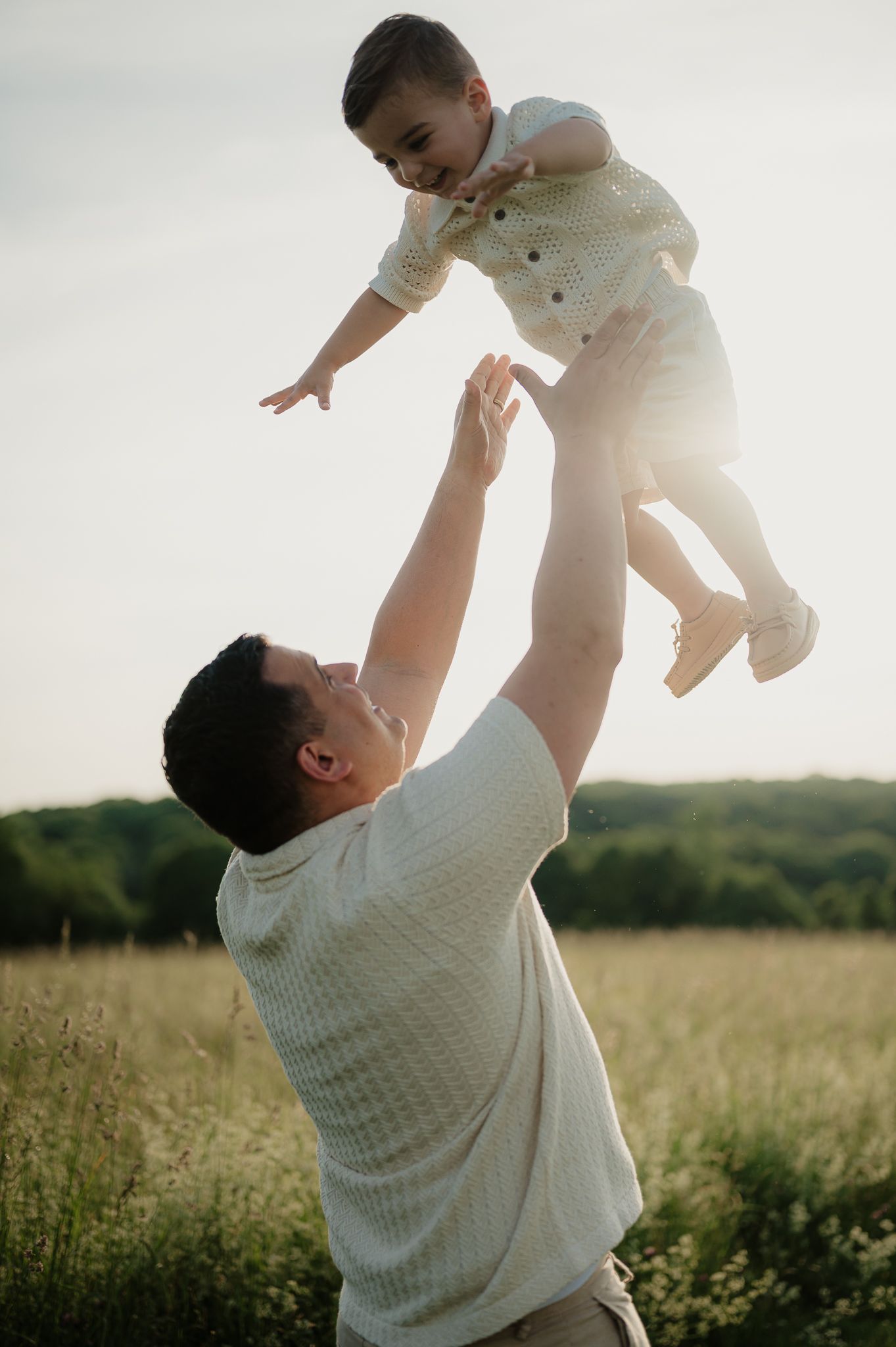 Father joyfully tosses young child in the air in a sunlit field, sharing a playful moment outdoors.