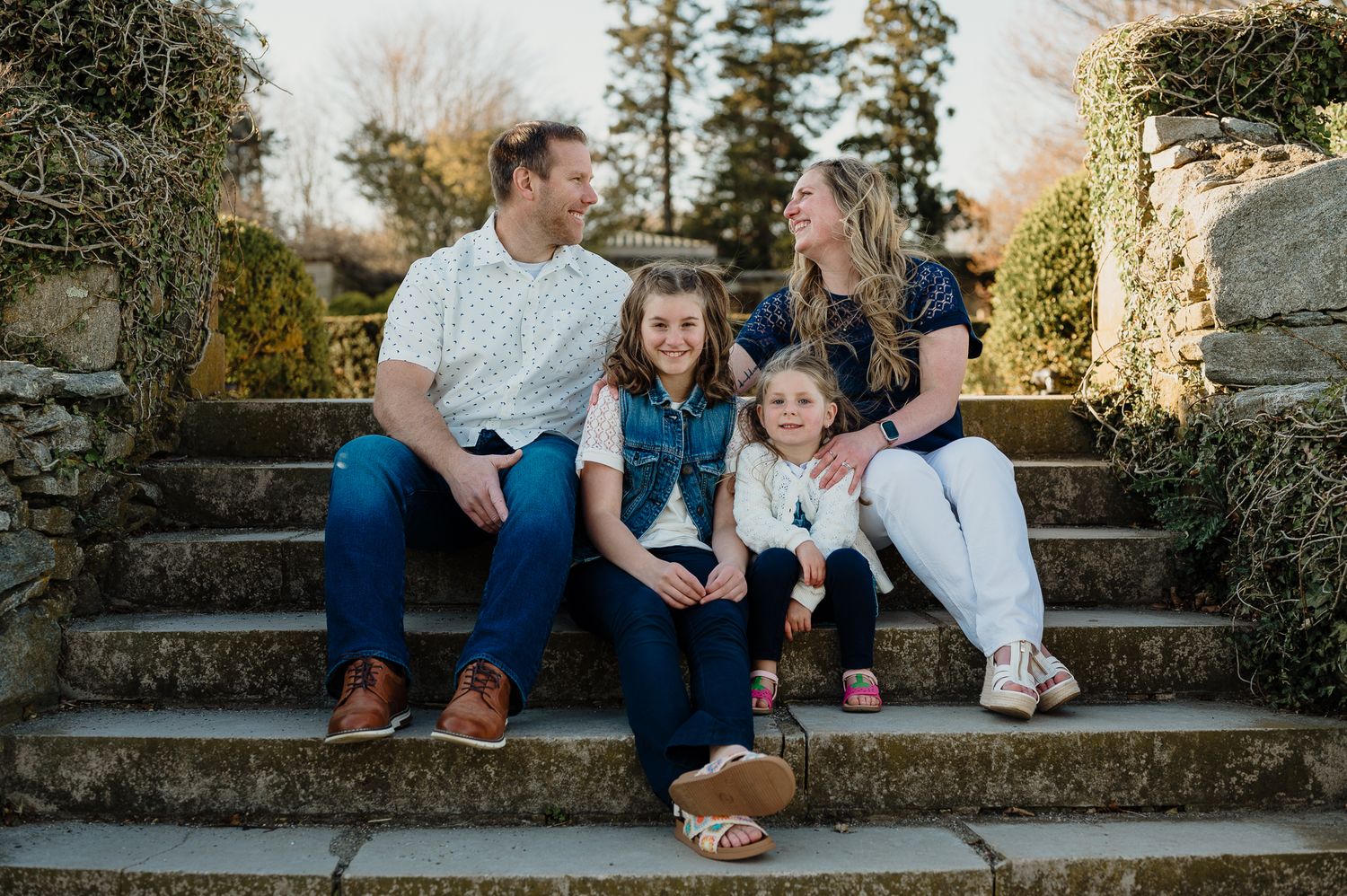 Happy family of four sitting on stone steps outdoors, smiling together in casual attire during golden hour.