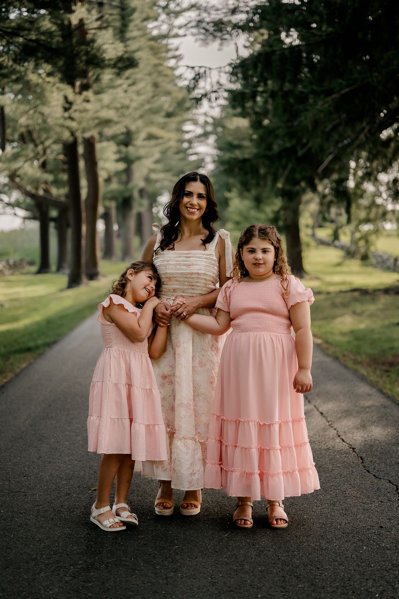 A woman in a floral dress stands with two girls in matching pink dresses on a path.