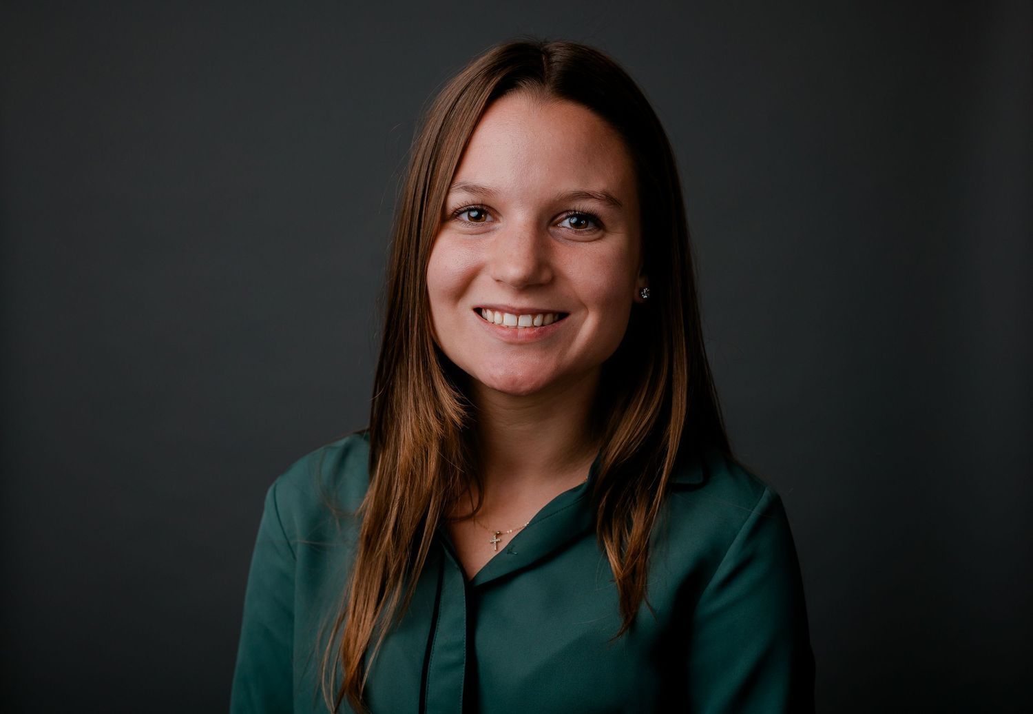 Professional headshot of a young woman with long brown hair wearing a green blouse on dark background.