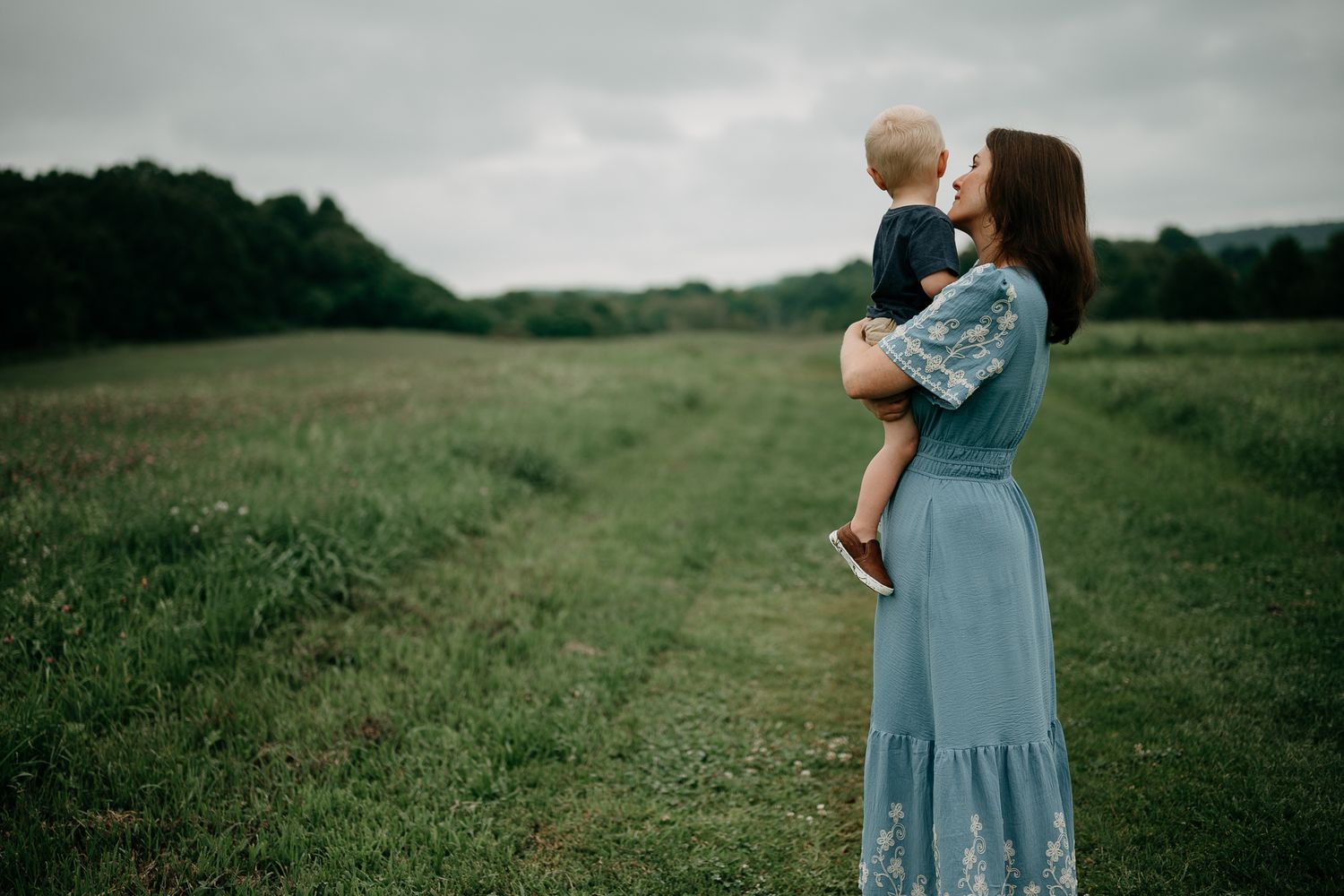 A mother in a blue floral dress holds her toddler in a lush green field under an overcast sky.