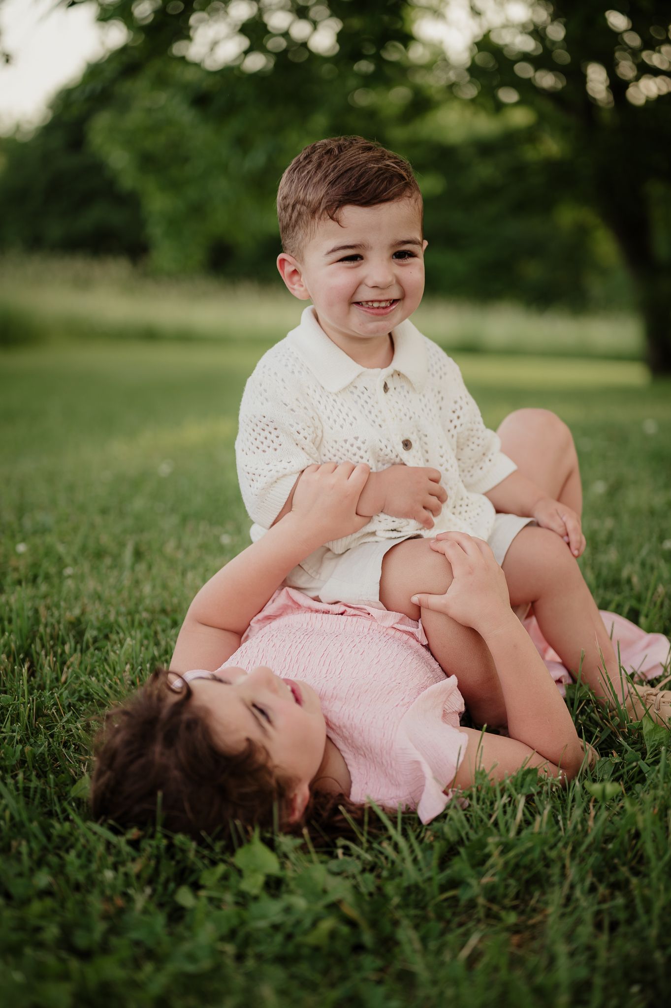Two young siblings playing on grass, boy in white knit top sitting with laughing girl in pink outfit.