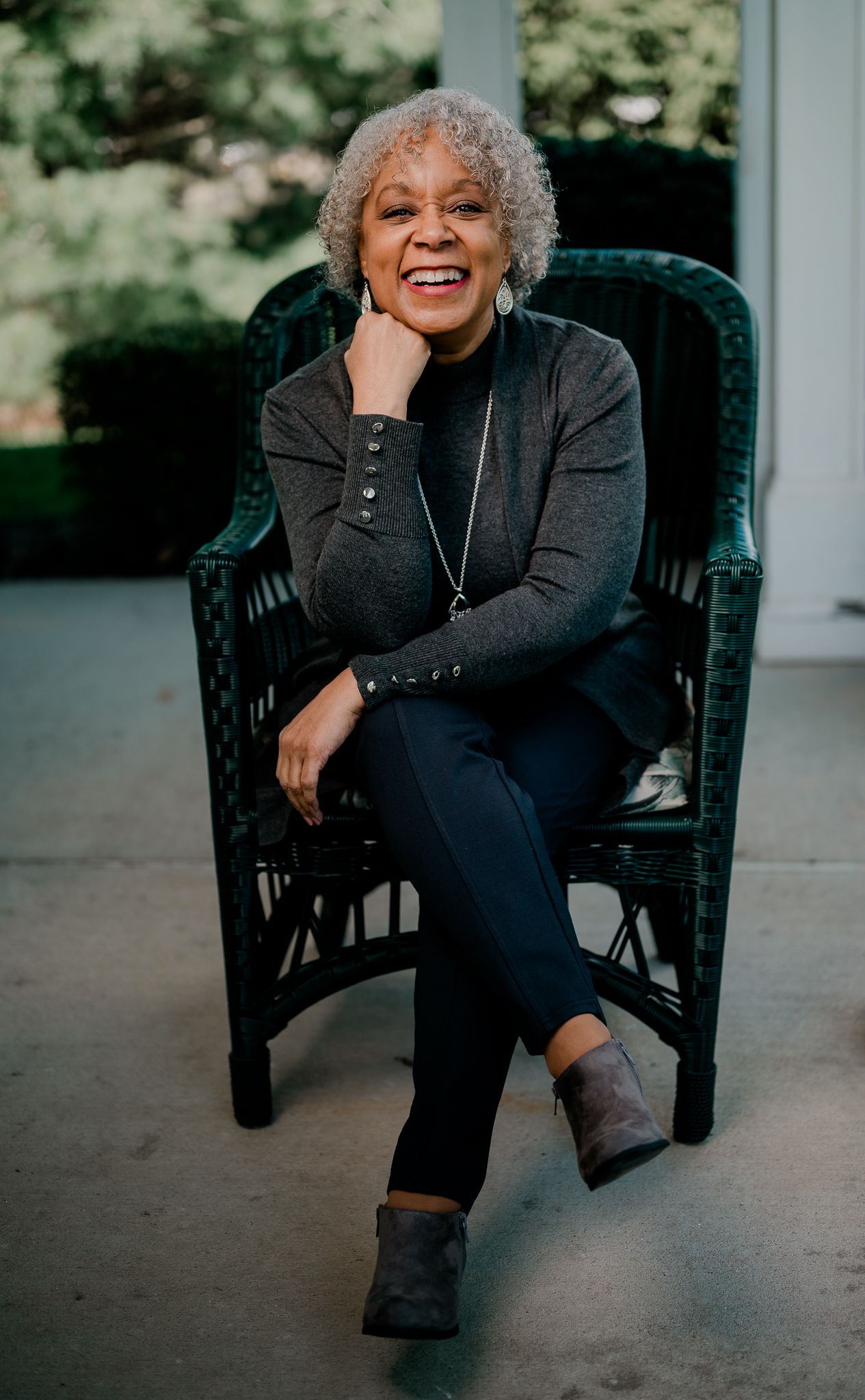 An older woman with gray curly hair laughs joyfully while seated in a dark wicker chair outdoors.