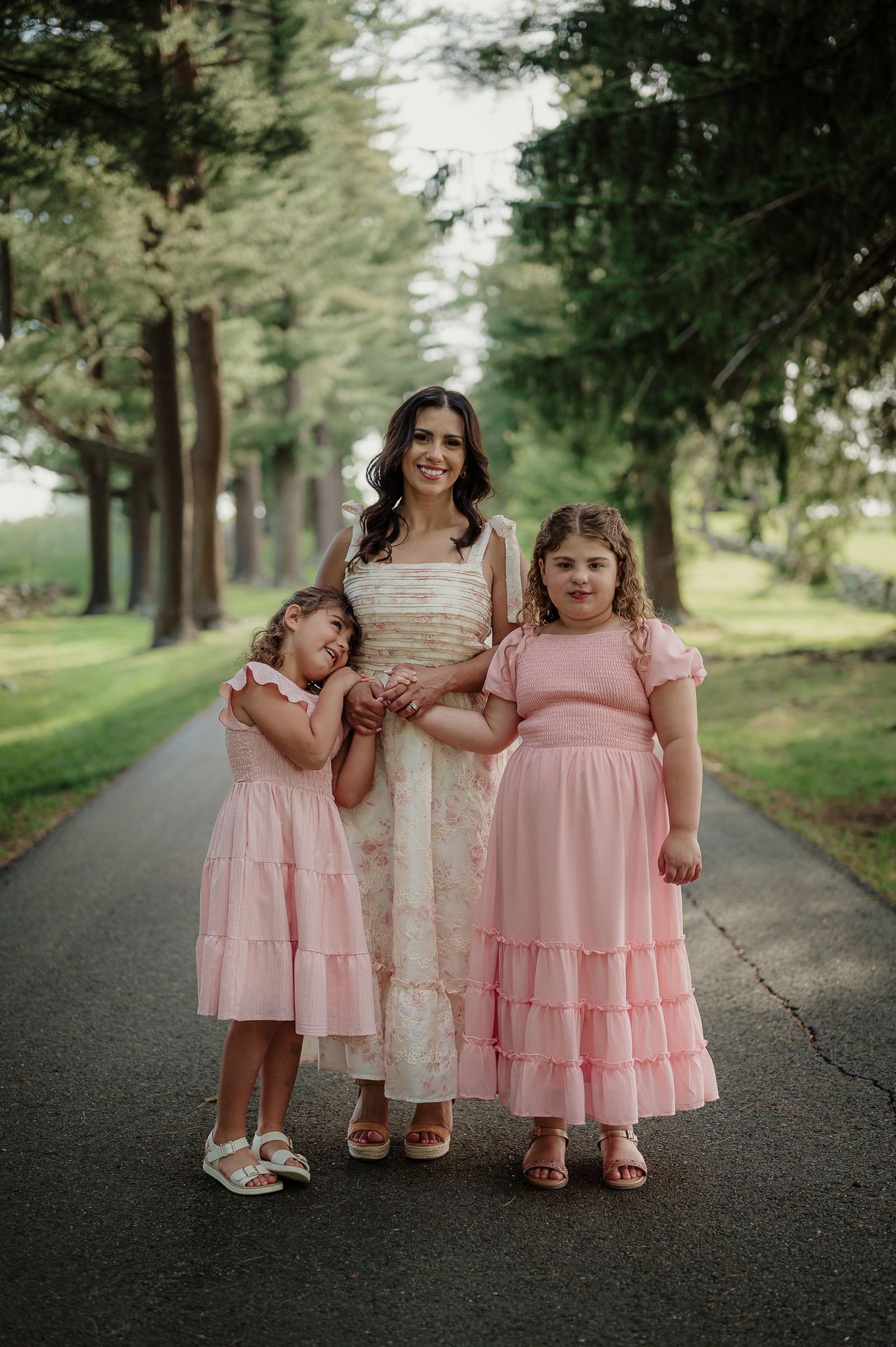 Woman in cream dress poses with two girls in pink dresses on a tree-lined path outdoors.