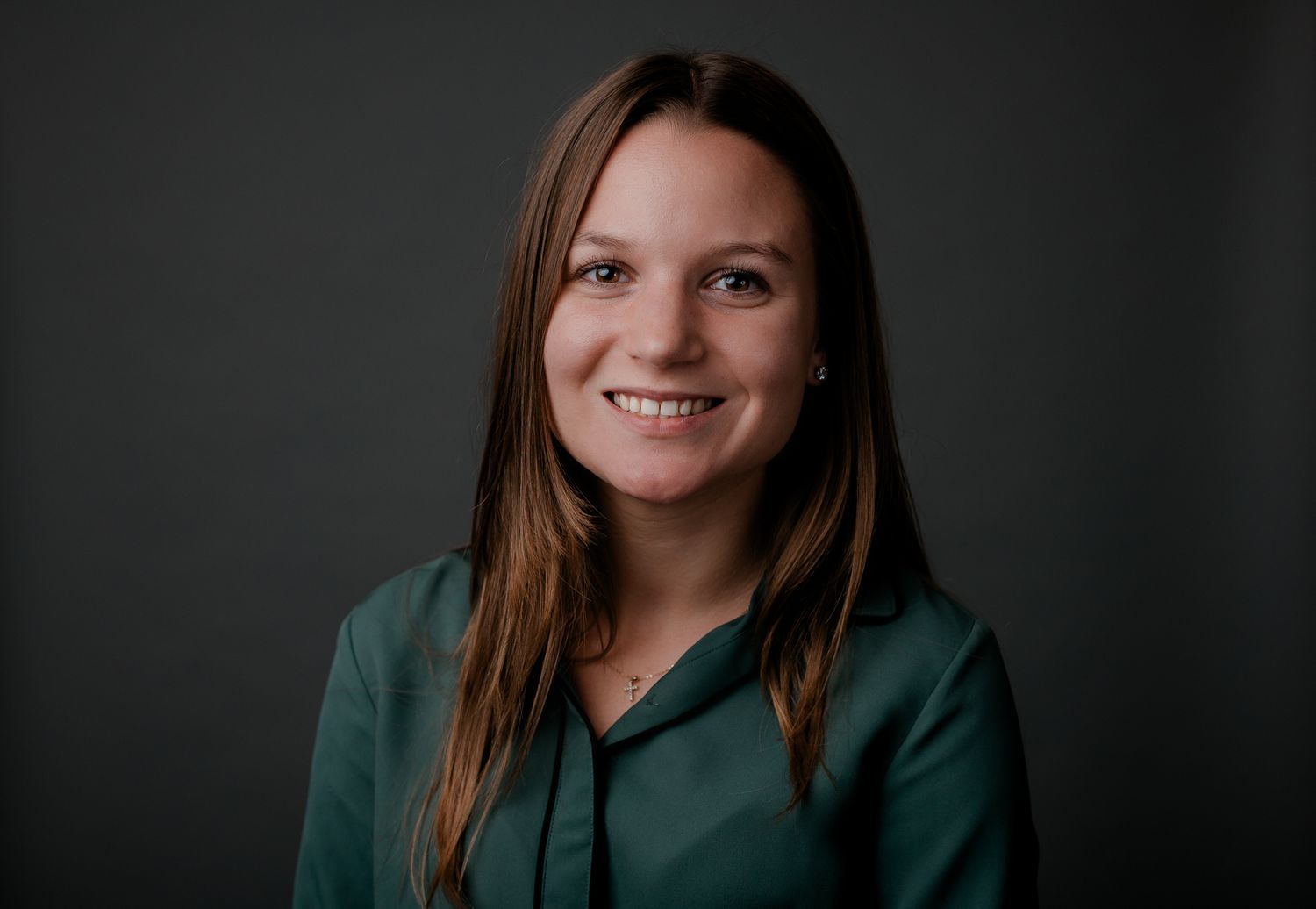 Professional headshot of smiling young woman in green blouse against dark gray background.