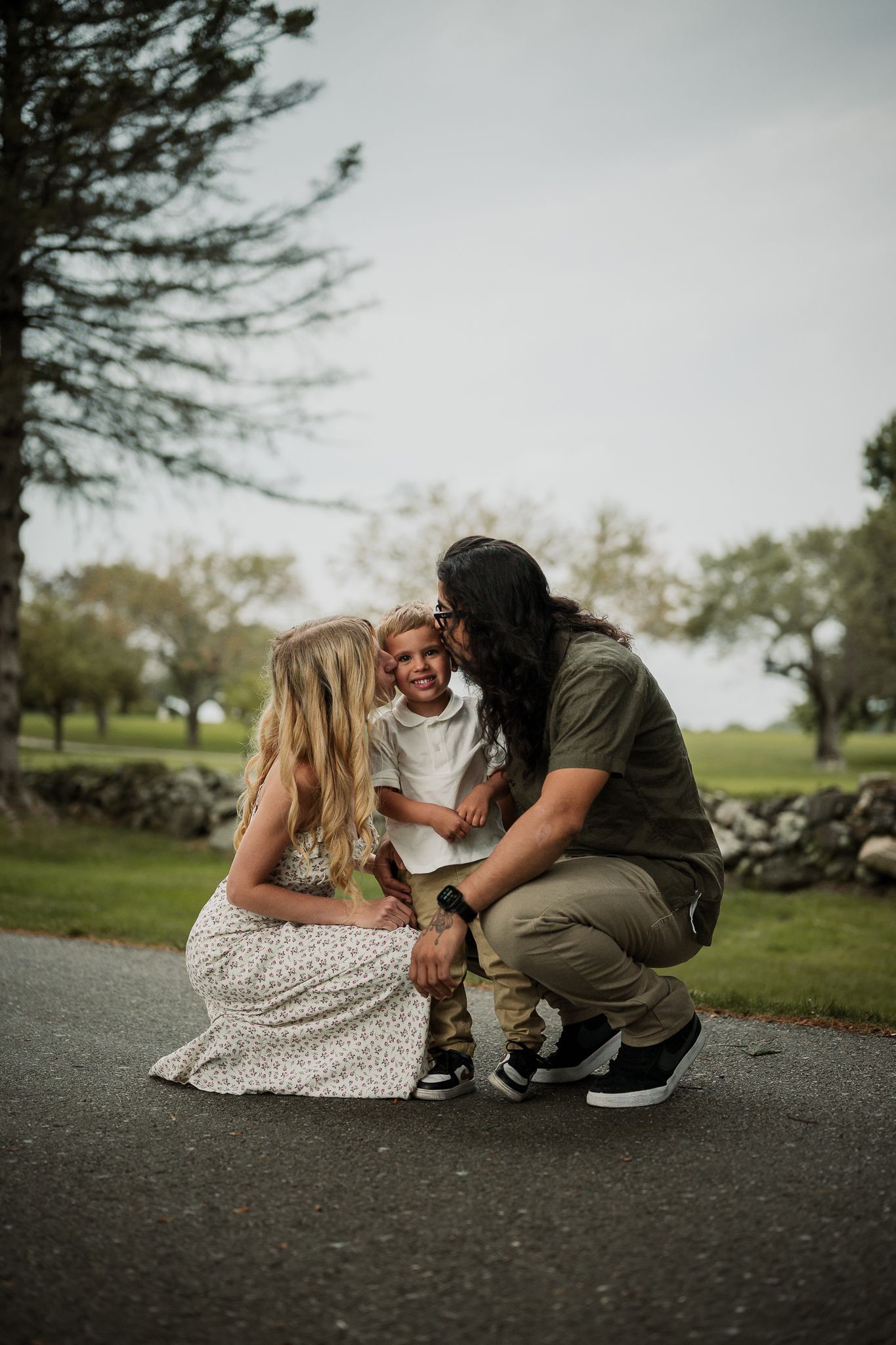 Family of three sharing a tender moment outdoors, two adults crouching to embrace a young child on a tree-lined path.