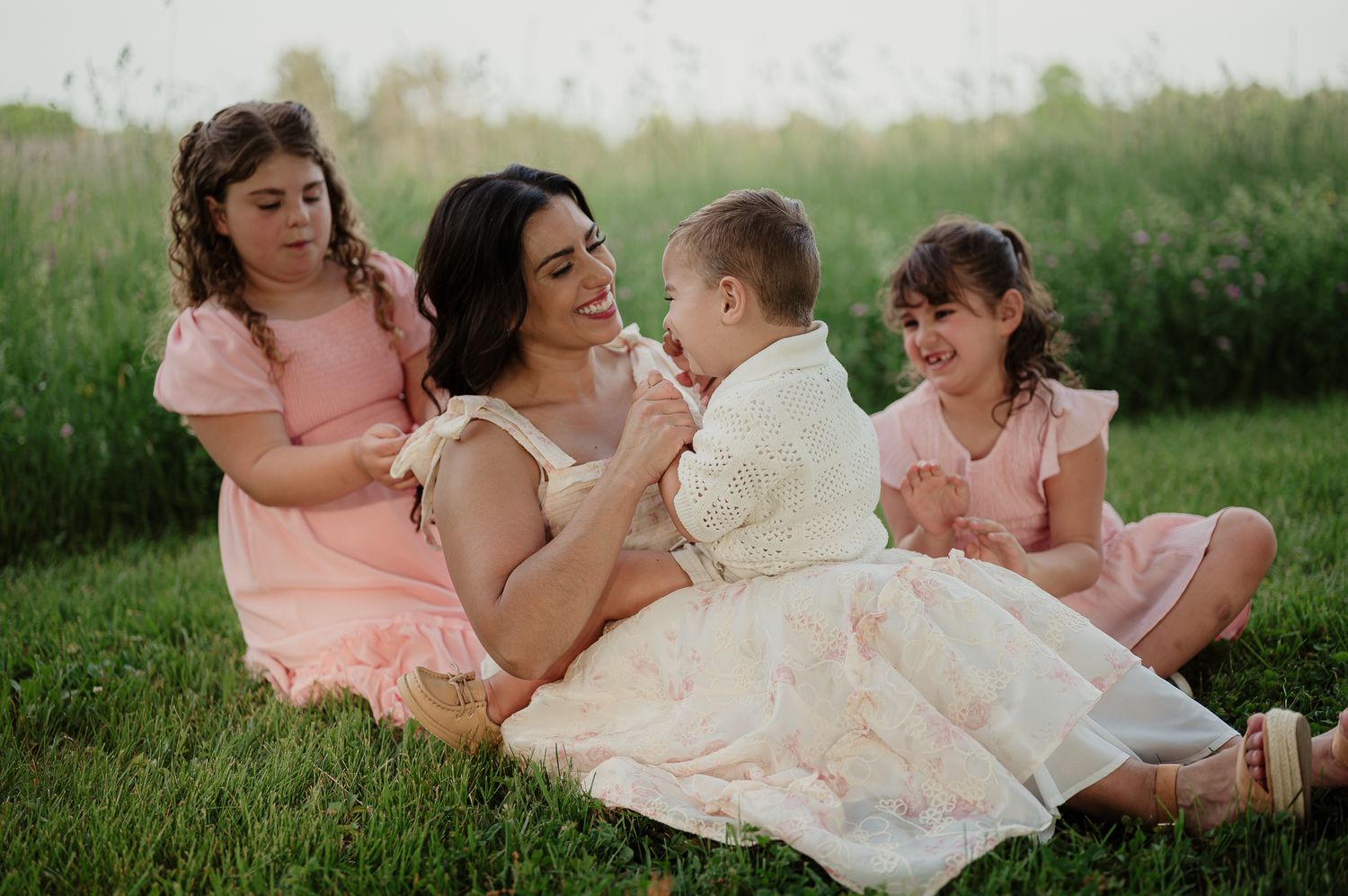 A mother sits on green grass with three young daughters in matching pink dresses during an outdoor family photo session.