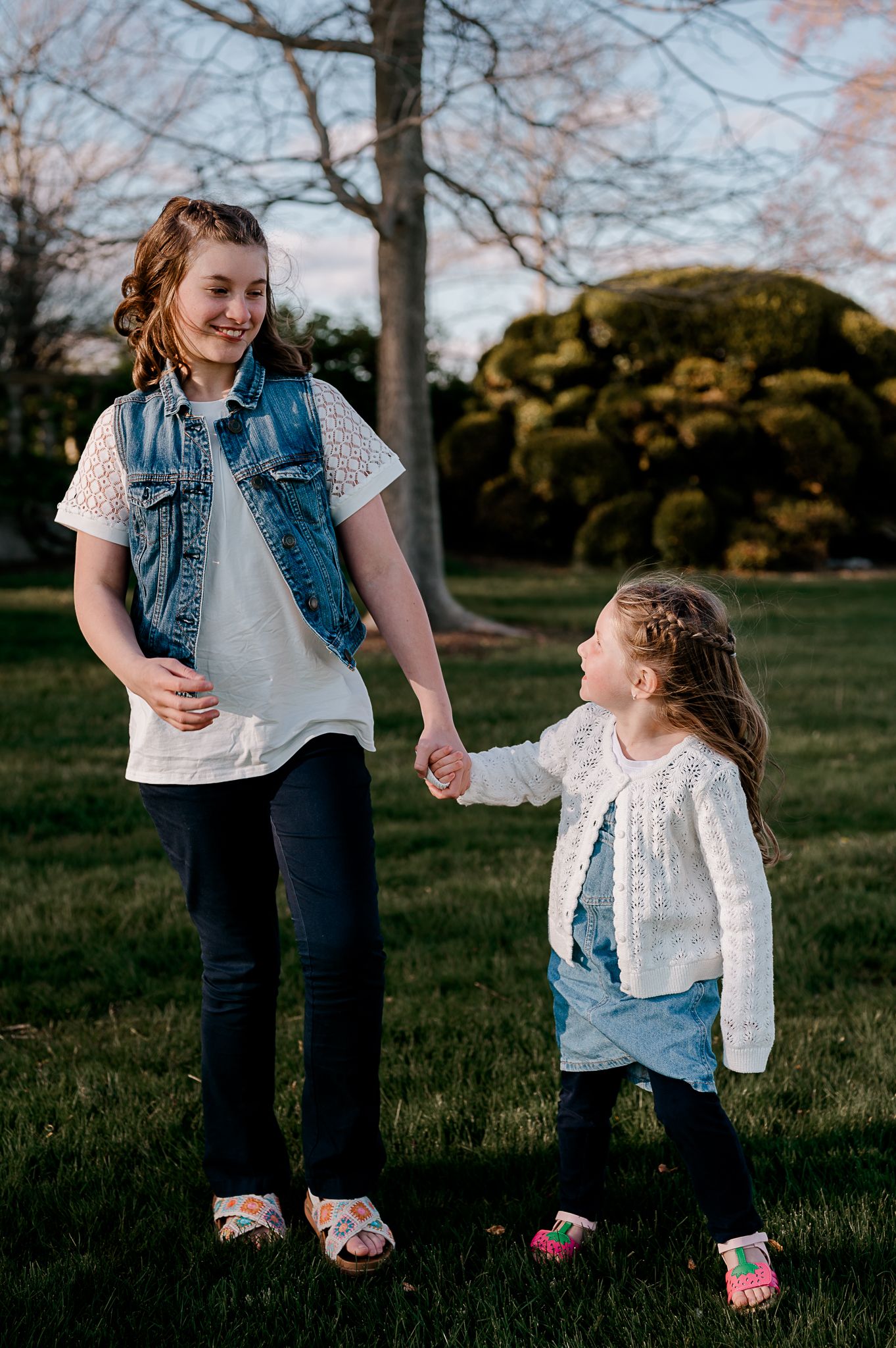 Two sisters holding hands and smiling in a park, older girl in denim vest, younger in white cardigan.