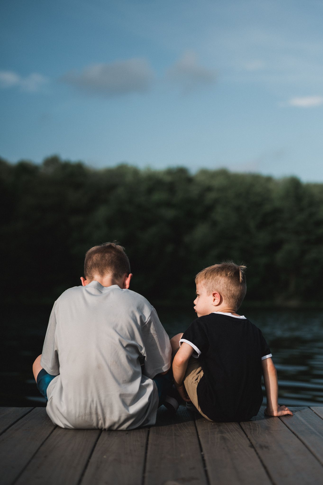 Two young boys sit together on a wooden dock overlooking a calm lake surrounded by lush green trees.