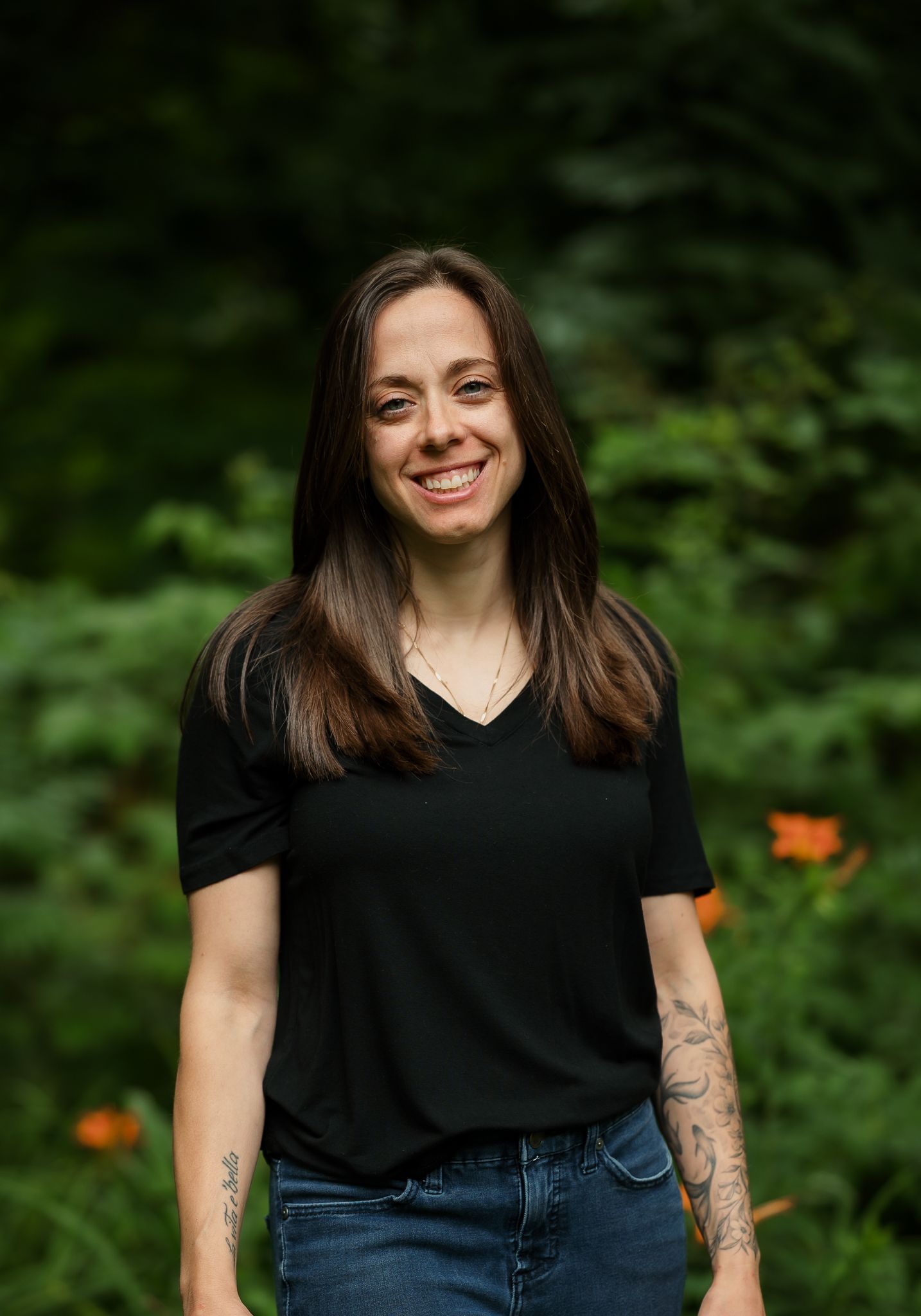 Smiling woman with tattoos wearing black t-shirt and jeans standing outdoors among green foliage.