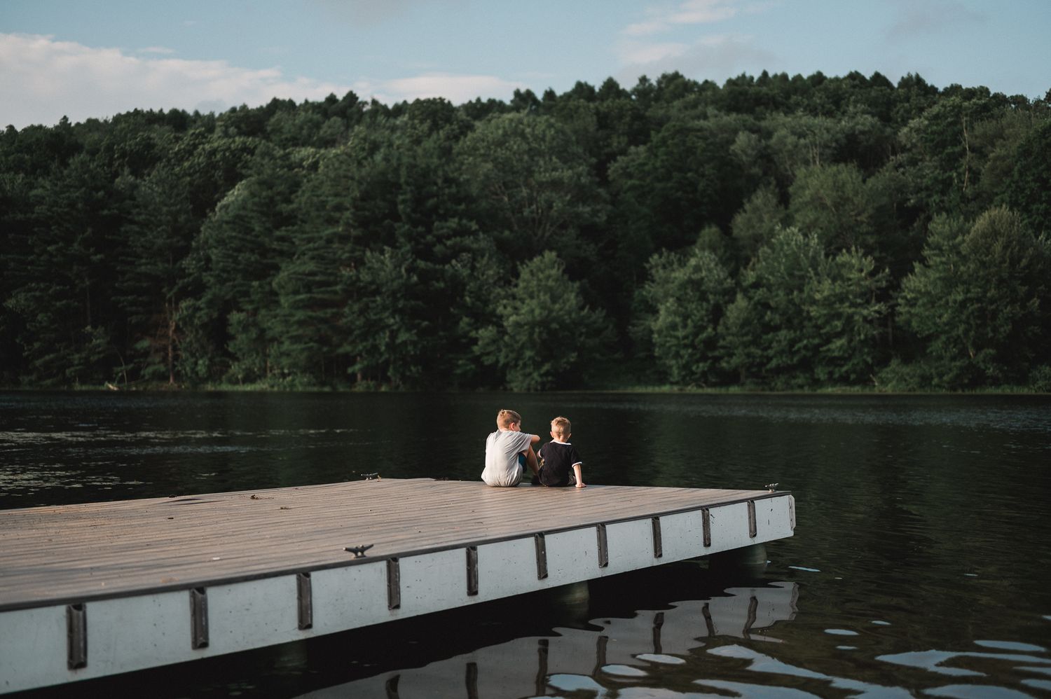 Two people sitting on a wooden dock by a calm lake surrounded by lush green forest trees.