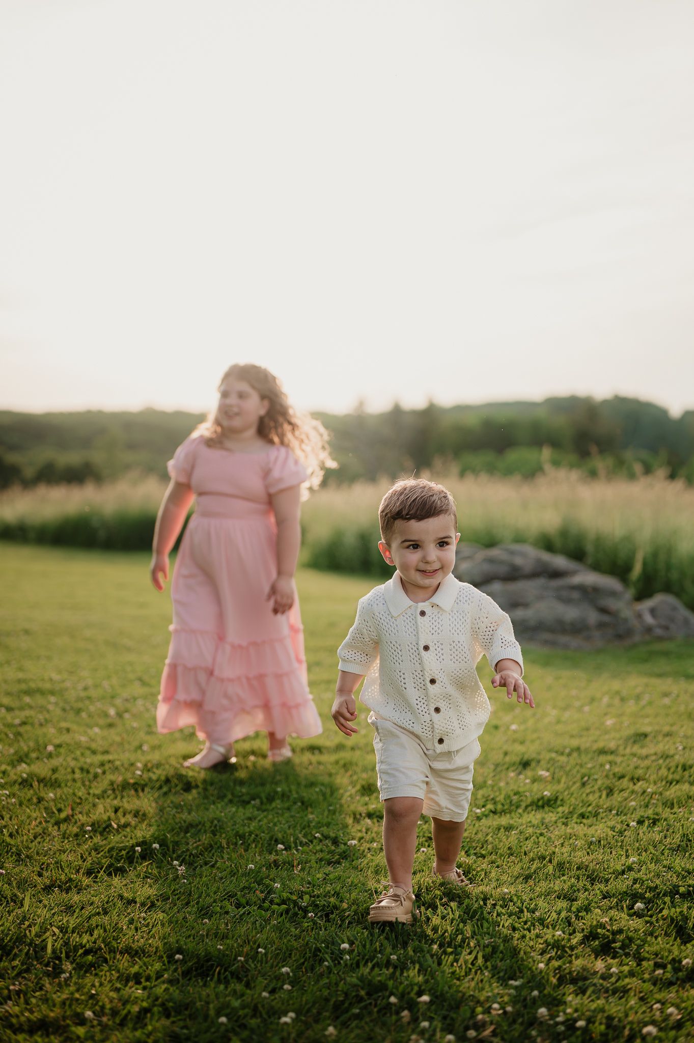 A young boy in a white outfit runs on green grass while a girl in a pink dress walks behind him at golden hour.
