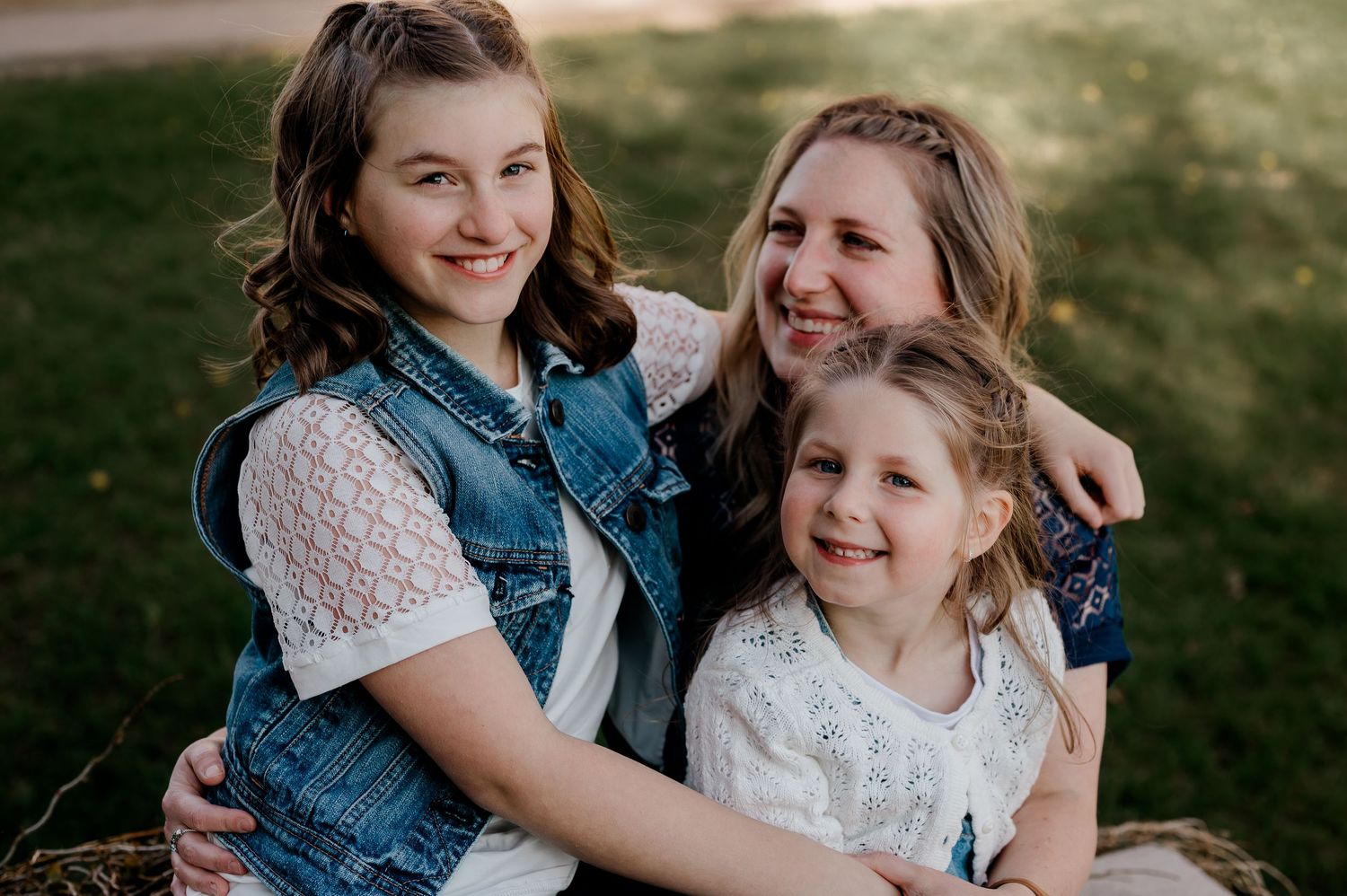 Three smiling girls sitting together outdoors on grass in casual denim and white outfits.