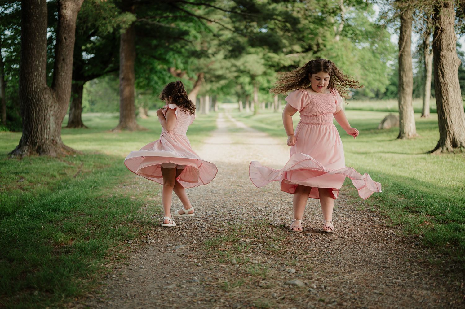 Two girls in pink dresses twirl on a tree-lined path in a lush green park.