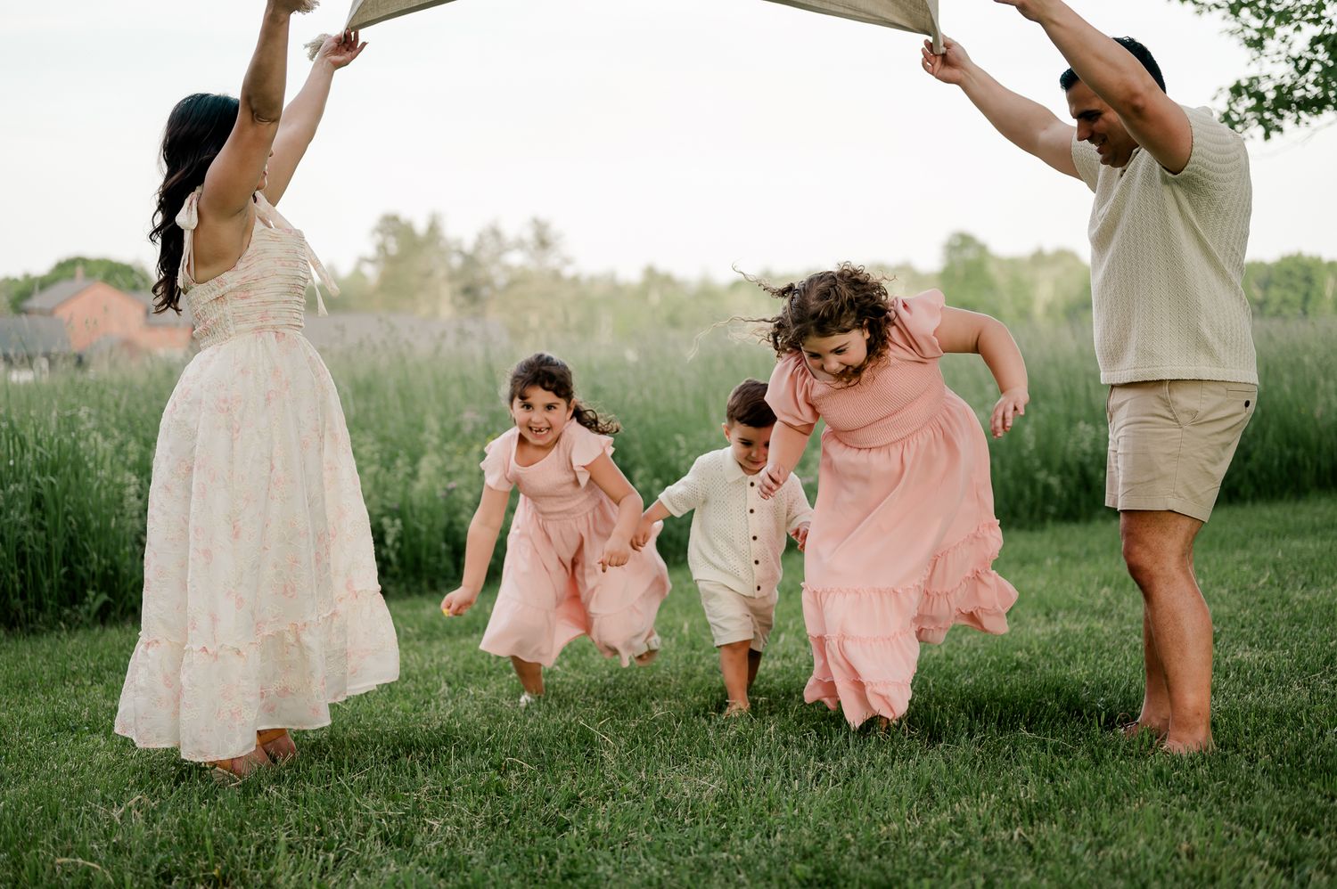 Happy family playing outdoors, parents lifting fabric arch while children run through on green grass.