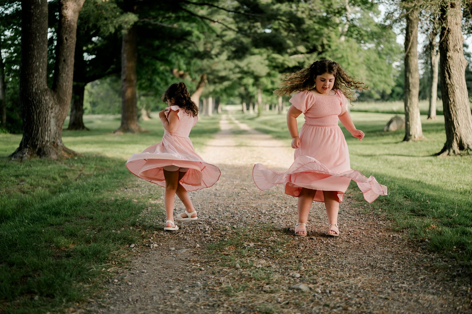 Two girls in pink dresses twirl and dance on a tree-lined gravel path outdoors.