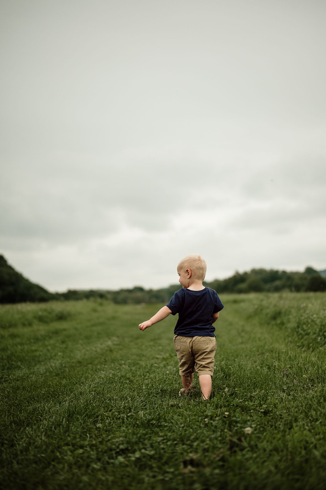 Young toddler boy walking alone through a lush green field on an overcast day, seen from behind.