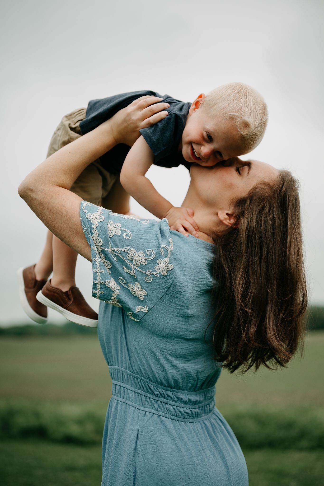 A mother in a blue dress joyfully lifts her smiling toddler son overhead in a green field.