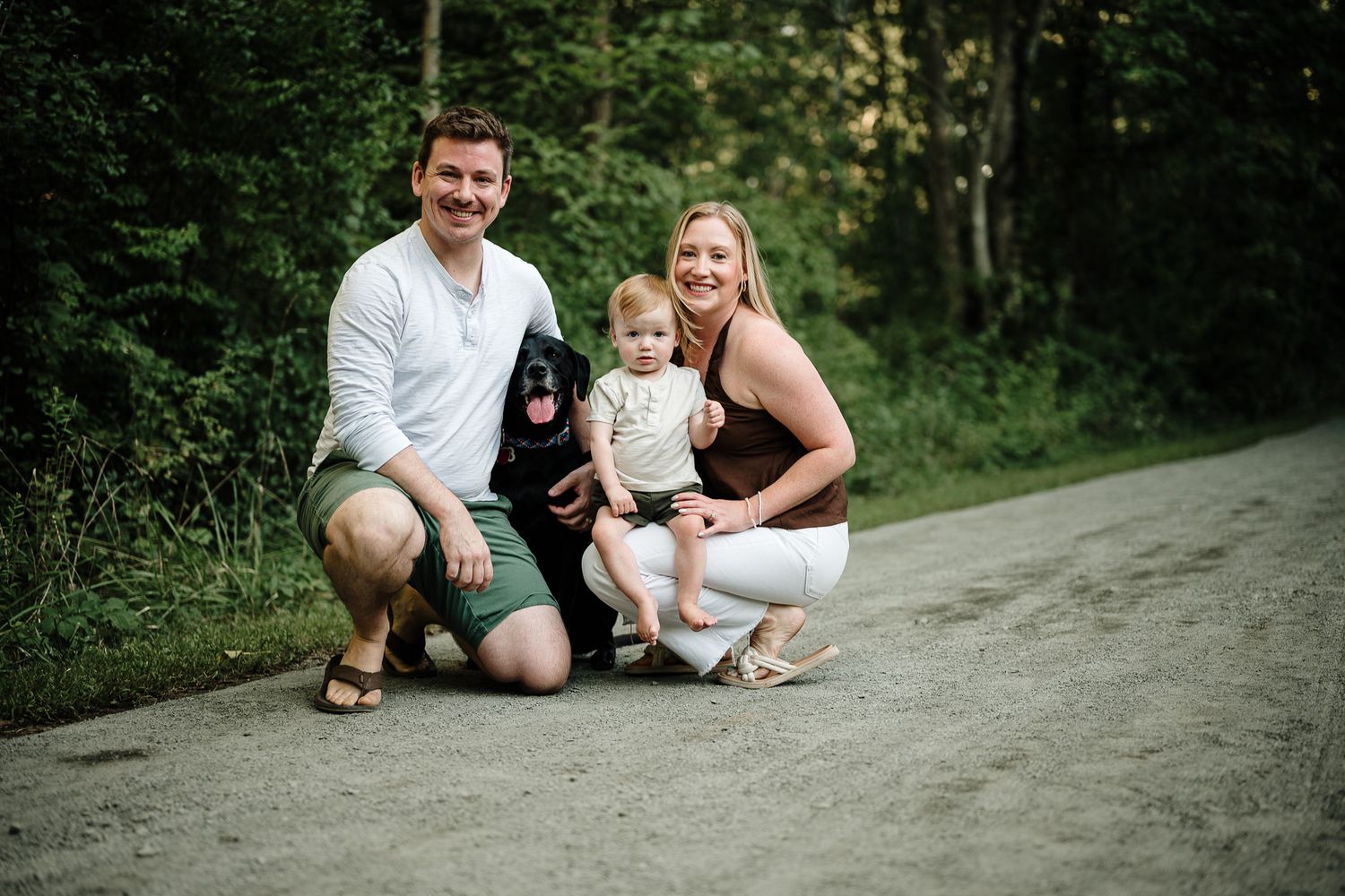 Happy family with baby and black dog posing on a dirt path surrounded by lush green forest.