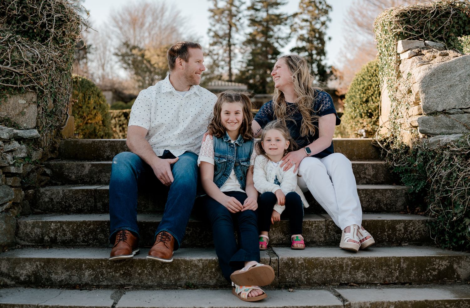 Happy family of four sitting on stone steps outdoors, smiling together in casual spring attire.
