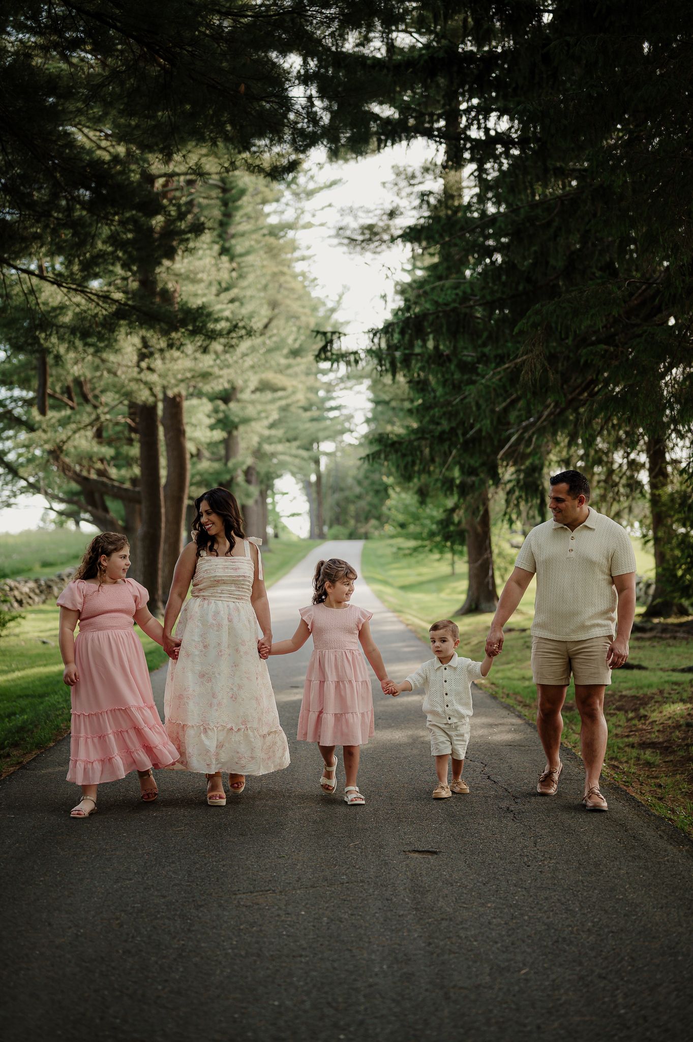 Family of five walking hand-in-hand down a tree-lined path, dressed in soft pink and neutral tones.